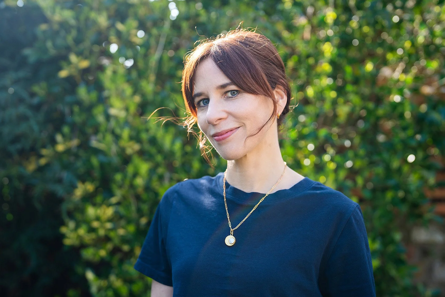 A woman with short brown hair and blue eyes, wearing a navy blue shirt and gold jewelry, standing outdoors in front of green foliage with sunlight backlighting her.