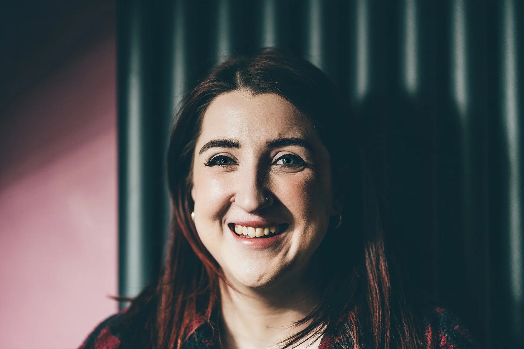 A woman with dark red hair smiling at the camera, standing in front of dark curtains and a pink wall.