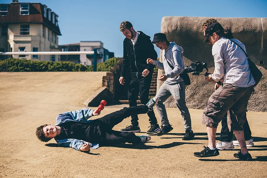 A young man on the ground pretending to be injured, while three individuals, including a photographer, are approaching him on a sunny outdoor location.