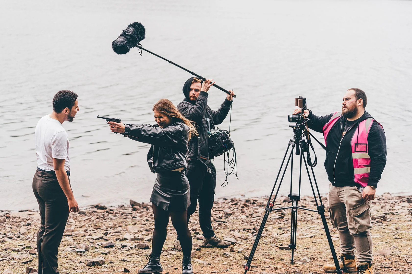 A film crew filming a scene at the shoreline with a woman aiming a gun at a man. The crew includes a cameraman, a sound operator holding a boom microphone, and a director or producer with a camera on a tripod. The scene takes place on a rocky beach with water in the background.