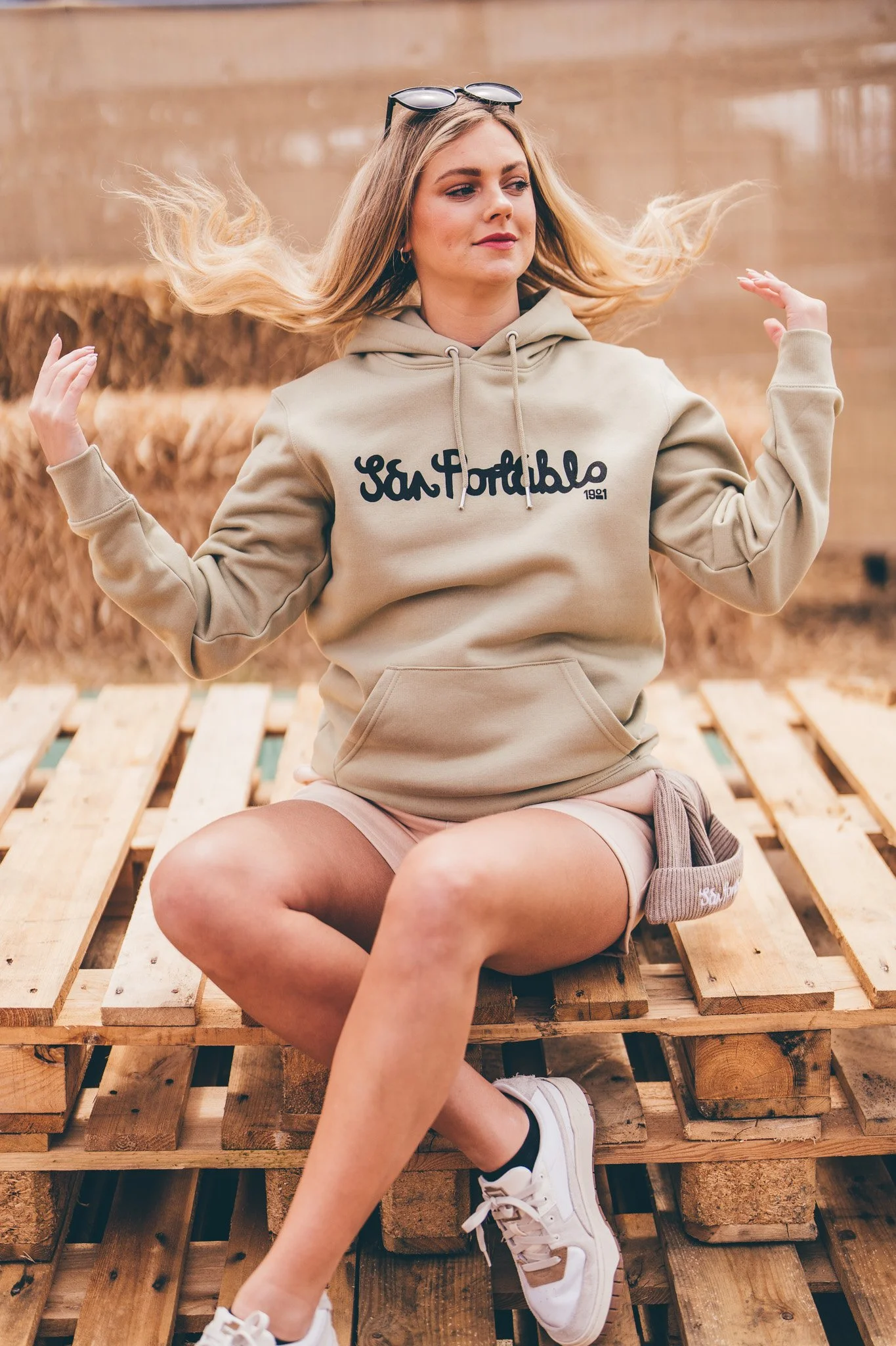 Young woman sitting on wooden pallets outdoors, wearing a beige hoodie with 'San Portfolio' written on it, shorts, sneakers, and sunglasses on her head, with her hair flowing in the wind.