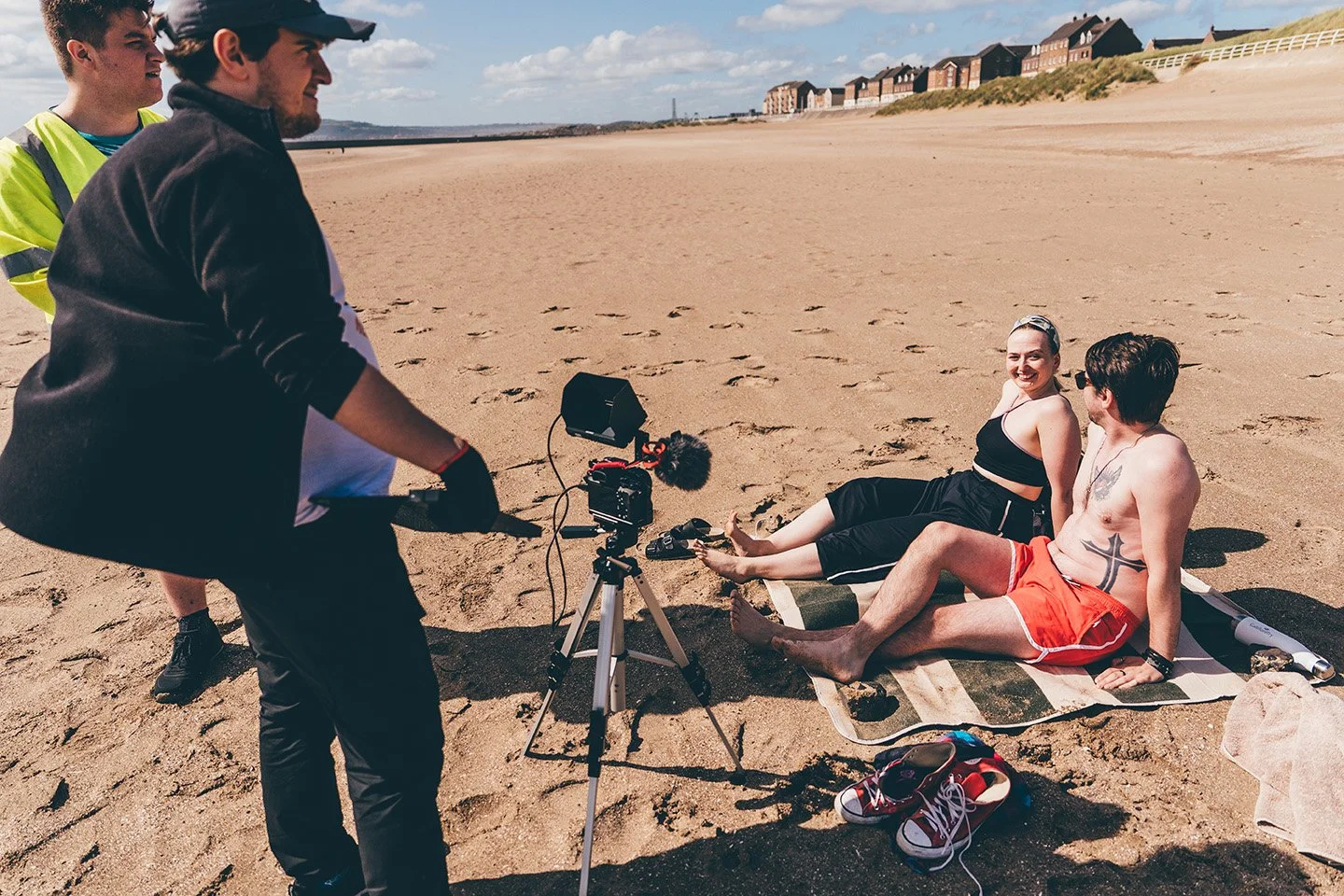 A film crew is recording two people sitting on a beach towel, a woman and a man with a tattoo, smiling at each other. The crew is using a camera mounted on a tripod, and the scene is set on a sandy beach with a row of houses in the background under a