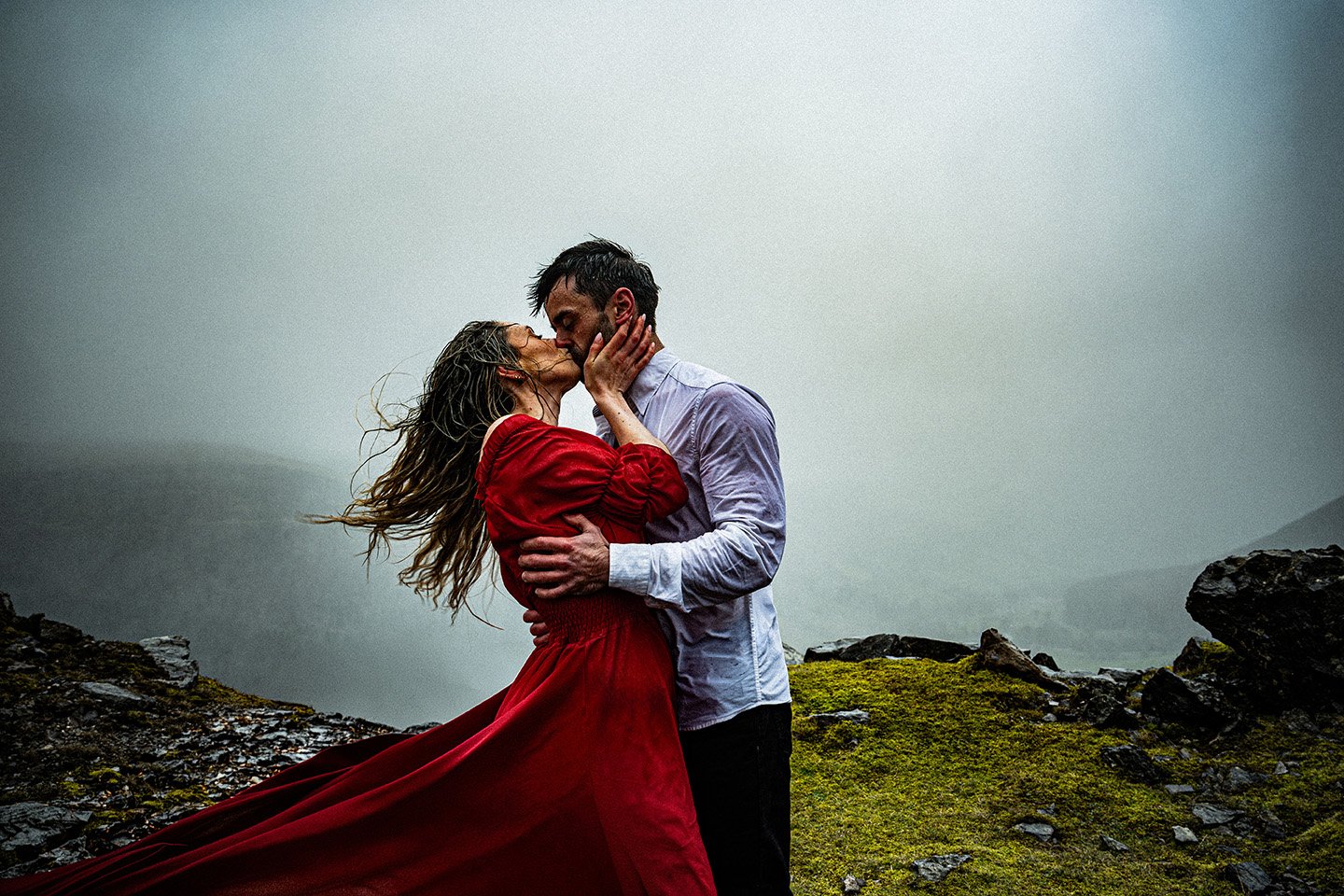 A couple embracing outdoors on a misty, rocky landscape, sharing a kiss.