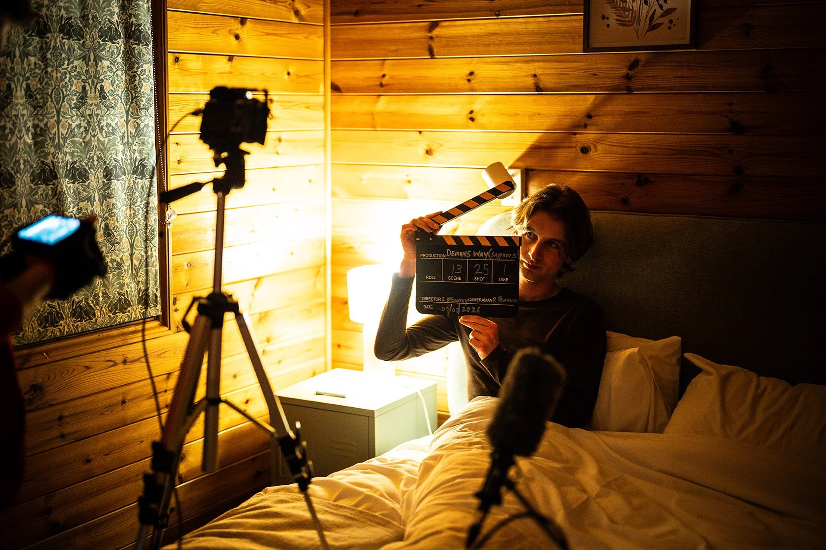 A film director holding a clapperboard while being filmed in a cozy room with wooden walls and warm lighting.