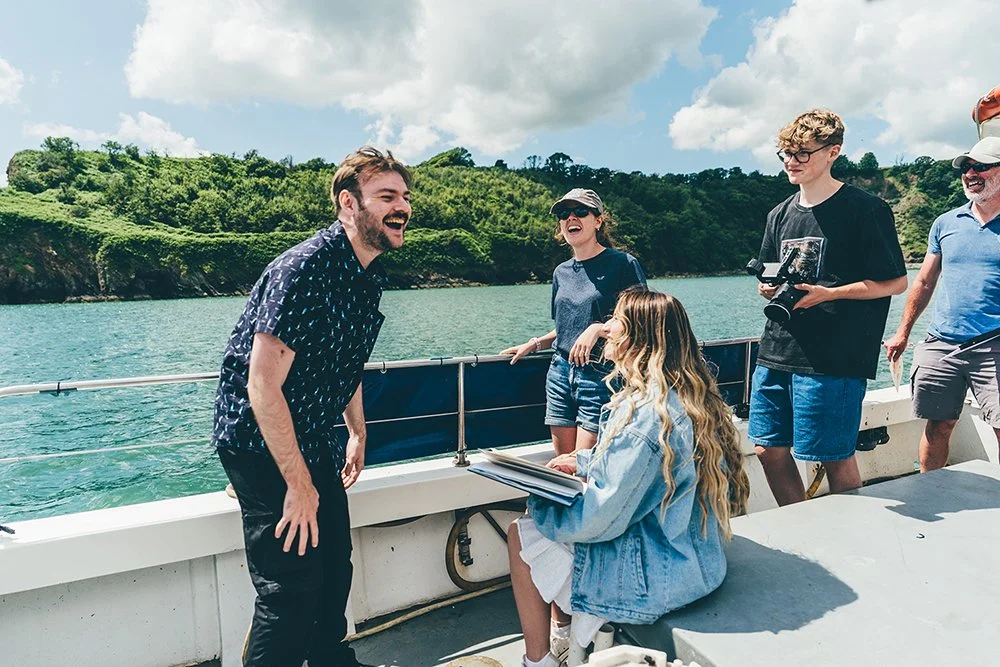 A group of people on a boat enjoying a day out, with one person laughing and others smiling and holding a drone controller, while a woman with long blonde hair, sitting and holding a book or notebook, looks up at him.