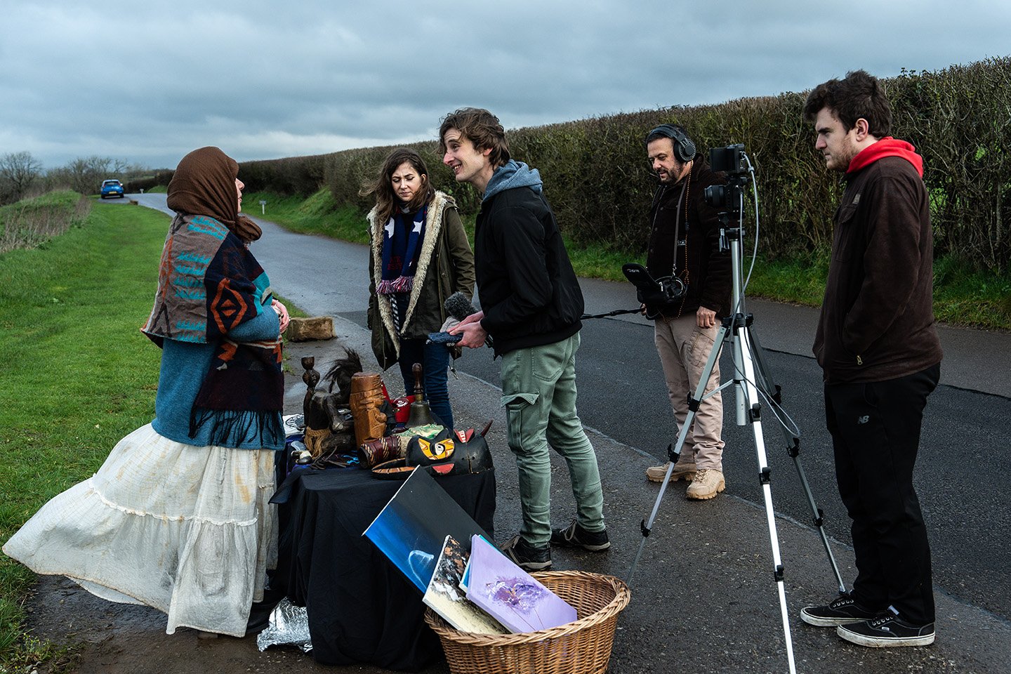 A woman dressed in a colorful jacket and long skirt meets a group of people filming her on a remote roadside surrounded by greenery and a hedge.