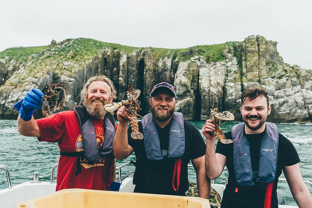 Three men on a boat holding live lobsters, with rocky green cliffs in the background.