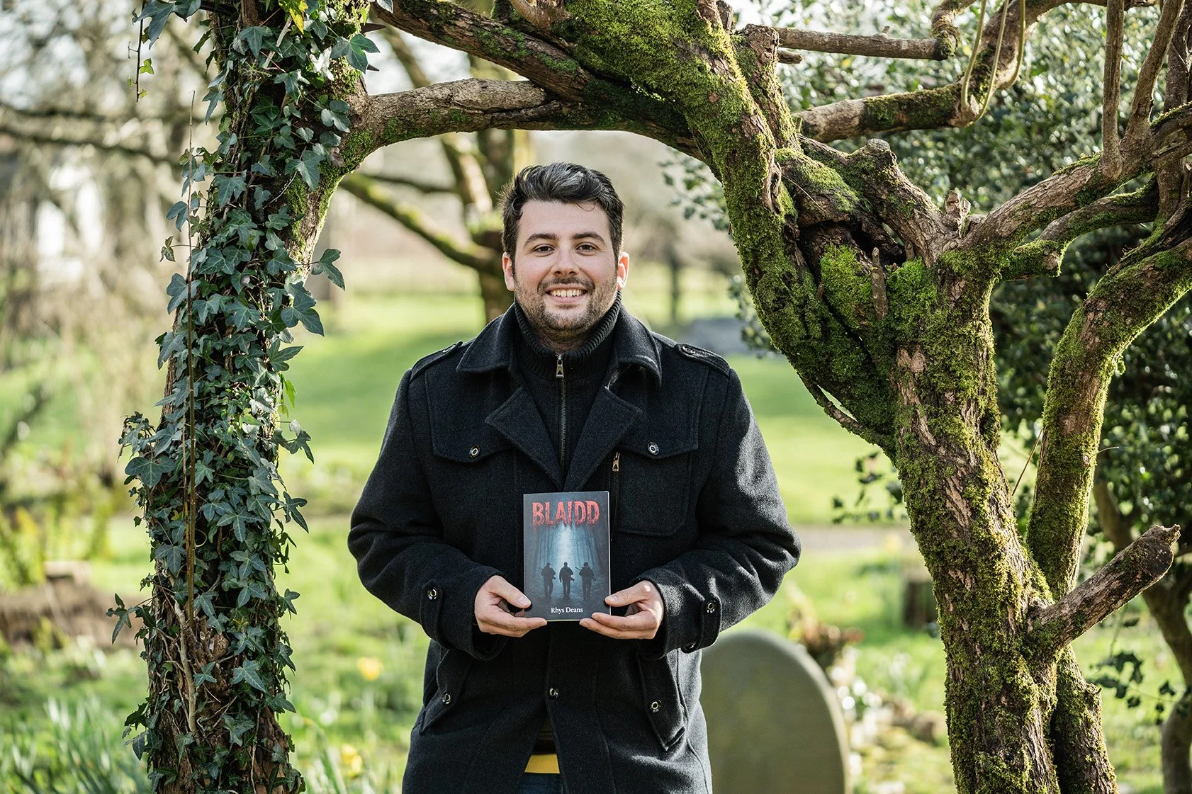 A smiling man holding a book titled 'BLAIDD' outdoors in a garden with moss-covered trees.