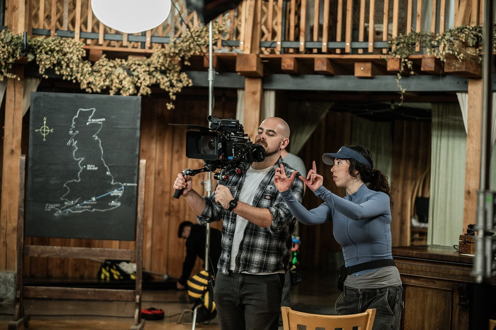 A man and woman working on a film set inside a wooden cabin or lodge. The man is operating a camera, and the woman is directing or giving instructions. There is a chalkboard with a map of the United Kingdom in the background and some decorations on the walls.