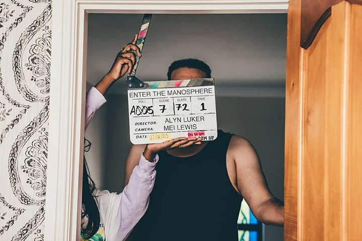 Person holding a clapperboard in front of their face, standing in a doorway, with another person's hand on a remote controlling the clapper. The clapperboard has text including 'Enter the Manosphere,' episode number, scene, slate, date, director's na