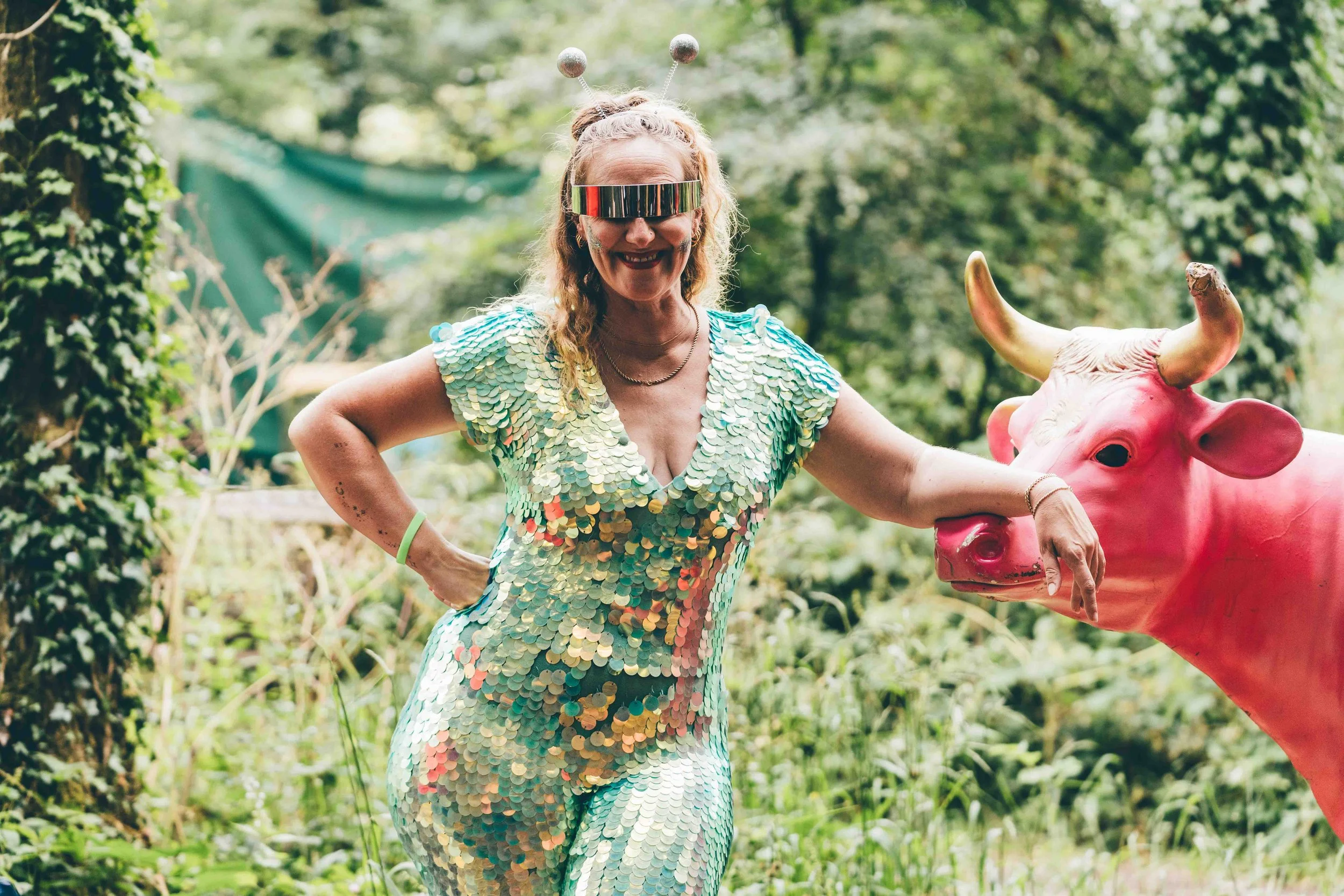 A woman dressed in a shiny, multicolored, sequin dress poses outdoors next to a pink cow statue. She is wearing a headband with antennae, sunglasses, and has a smile on her face.