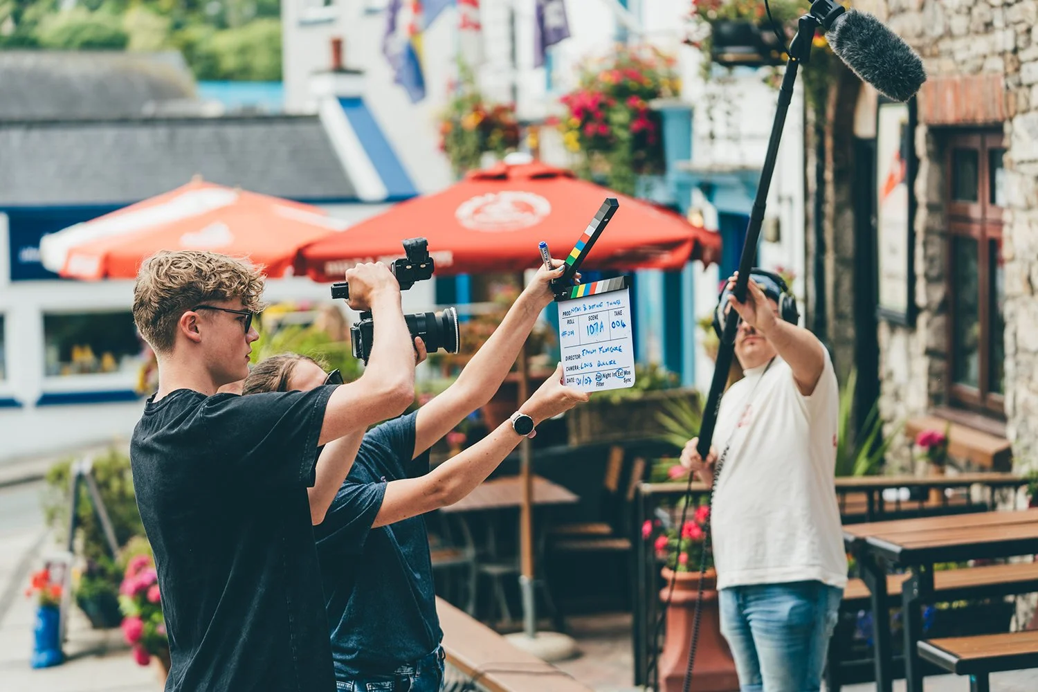 Film crew on outdoor set with a person holding a boom microphone and a woman holding a clapperboard during a film shoot.