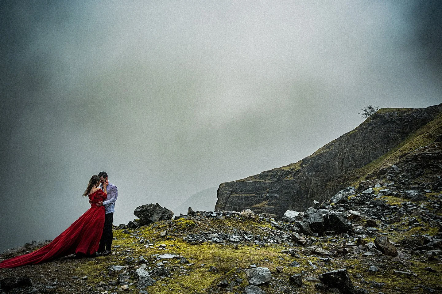 A couple embracing on a rocky, mossy landscape with a cloudy sky in the background.