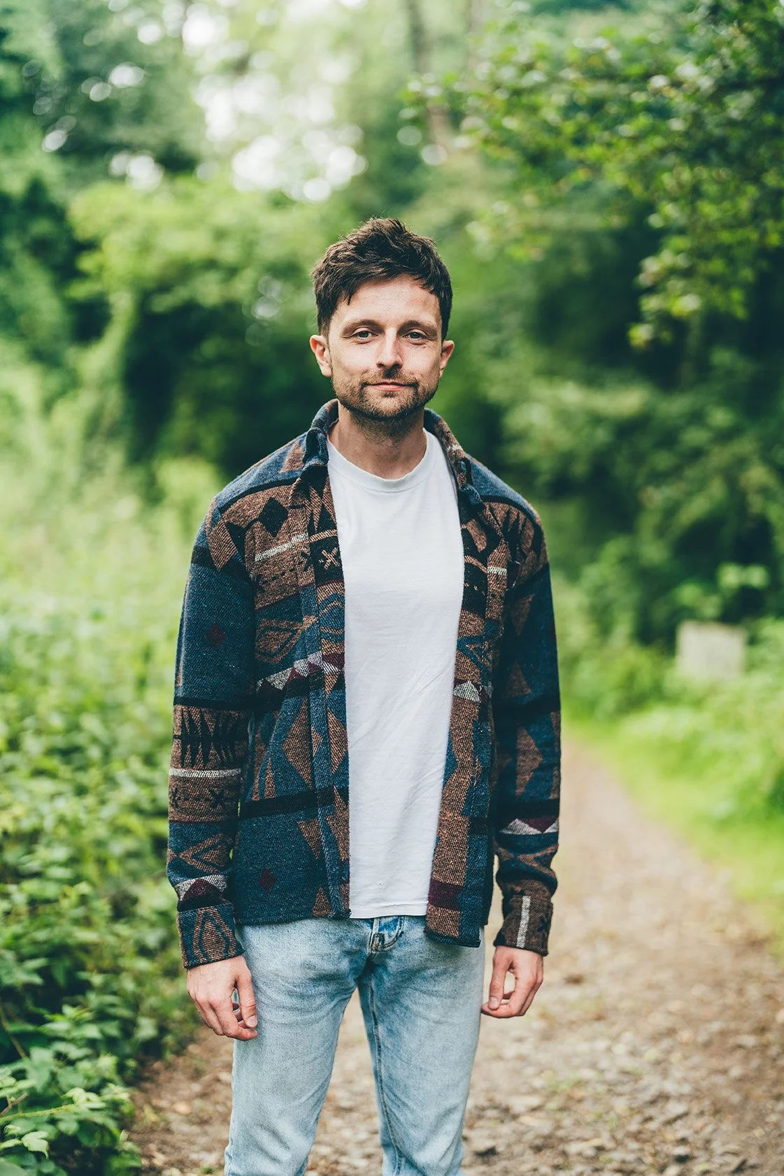 A man standing on a forest trail surrounded by greenery and trees, dressed in a patterned jacket, white t-shirt, and light jeans.