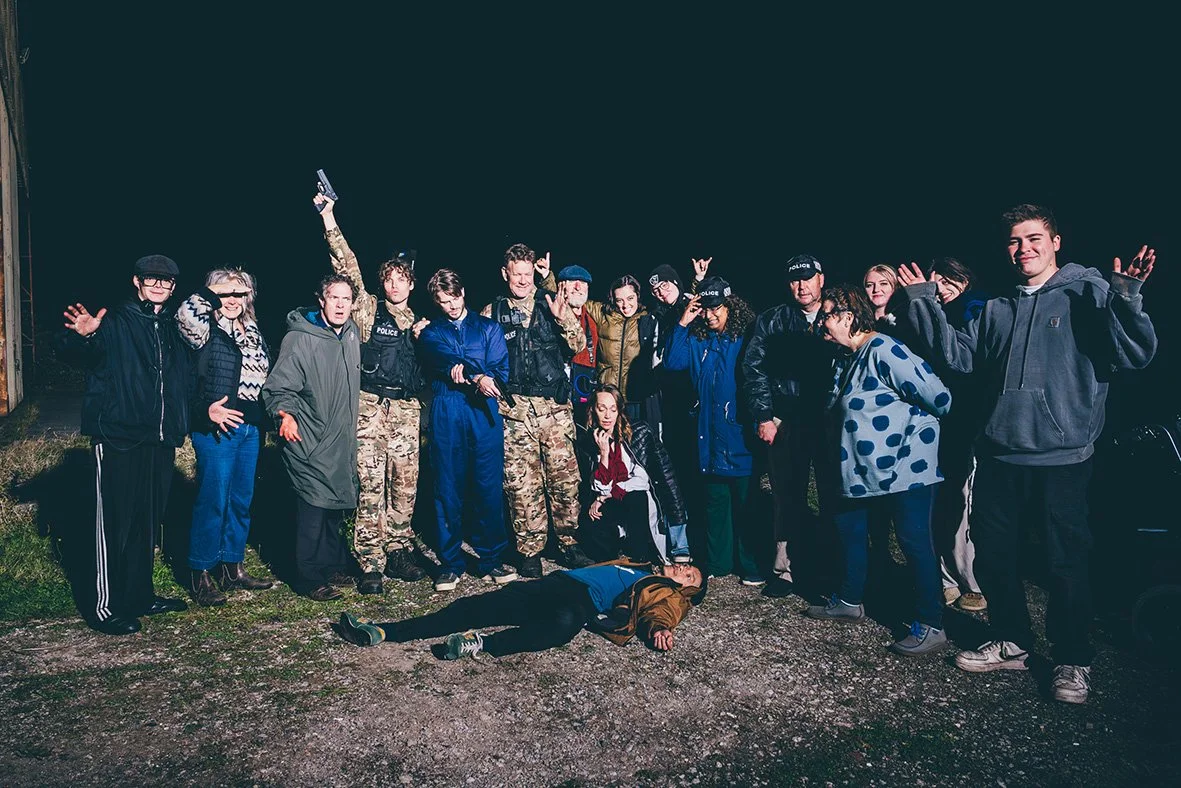 Group of people, including police officers and civilians, standing outdoors at night, with one person lying on the ground in front of them.