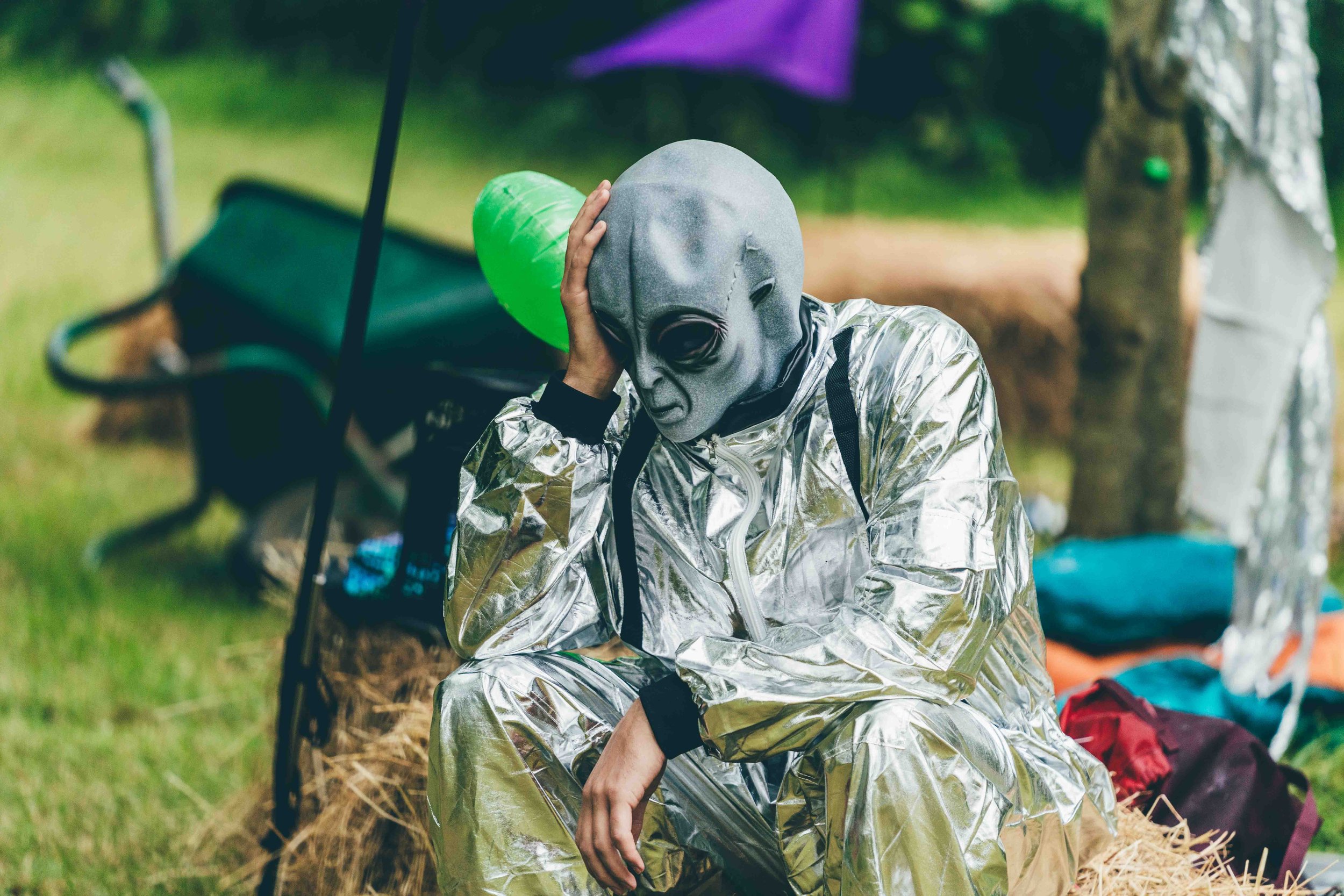 Person wearing a silver reflective suit and an alien mask, sitting on a hay bale outdoors, holding their head with one hand, with a green balloon attached to the mask.