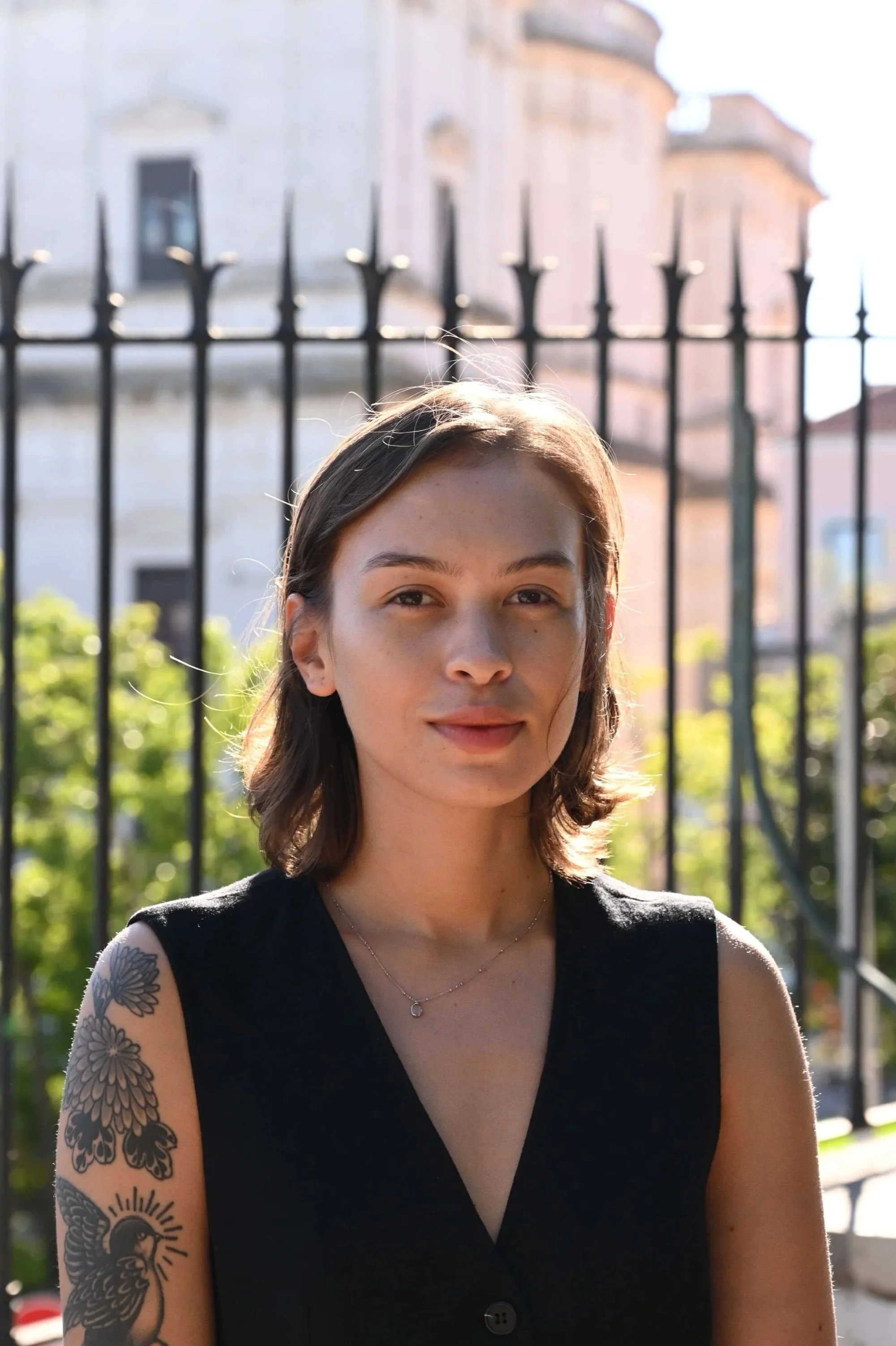 A young woman with shoulder-length brown hair, wearing a black sleeveless top, outdoors with sunlight illuminating her face and a tattoo on her left arm. Background shows a black iron fence, trees, and buildings.