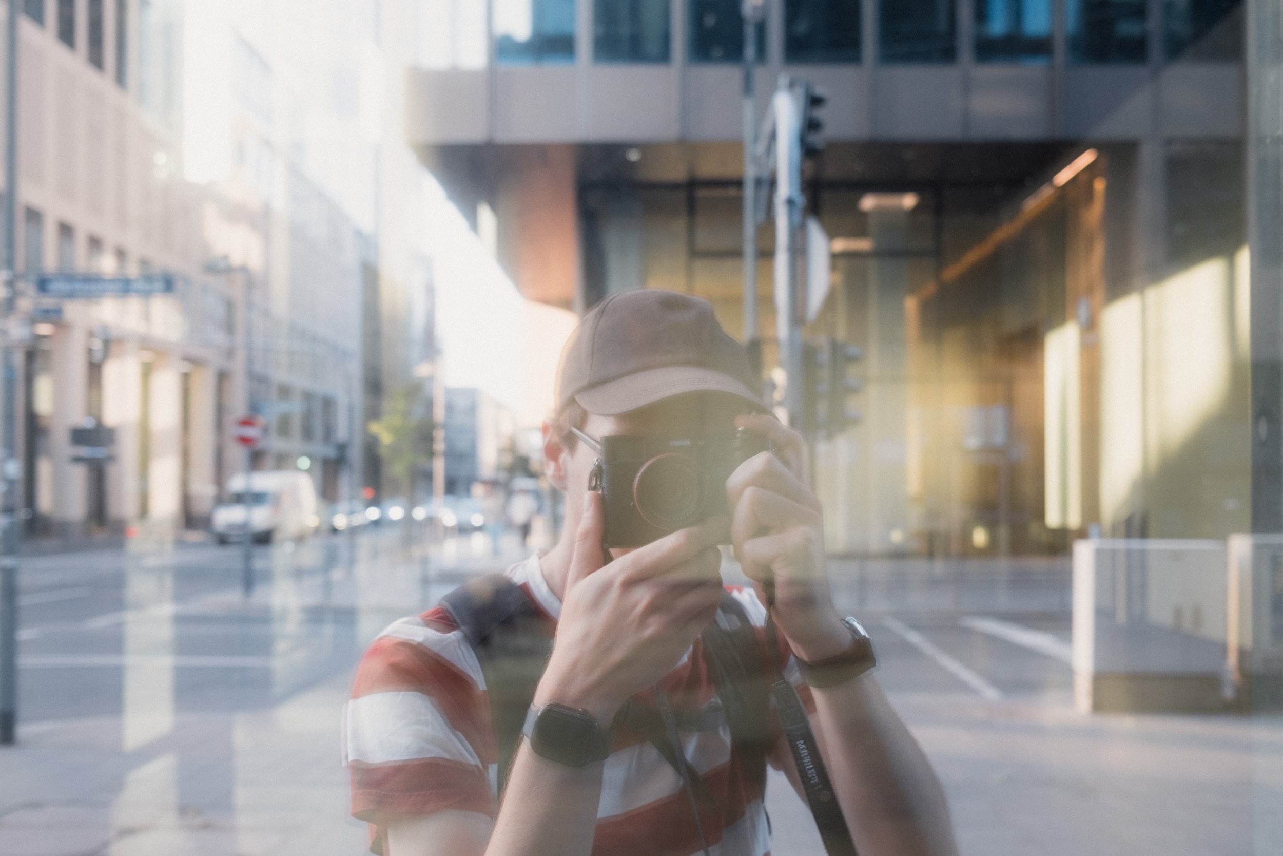 Reflection of a person taking a photograph with a camera, visible on a glass surface with city buildings and streets in the background.