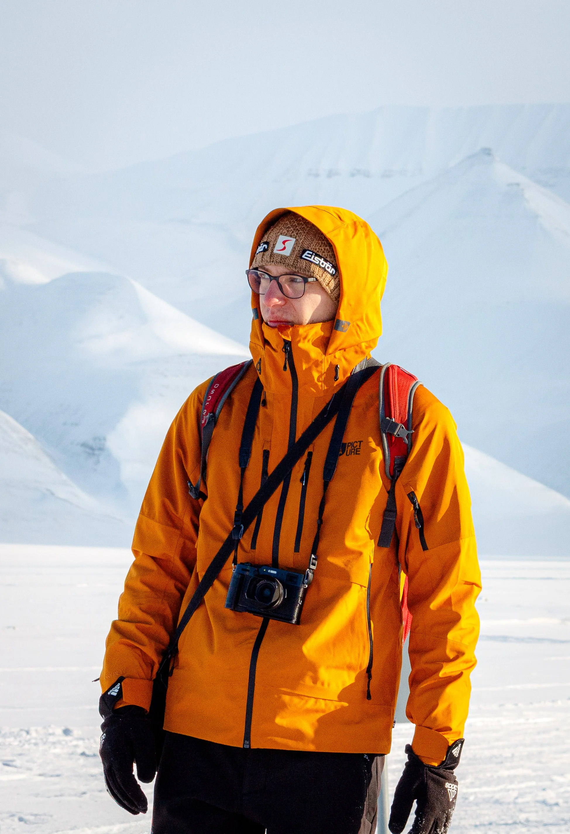 Person wearing an orange jacket and glasses, standing in snowy landscape with a mountain backdrop, holding a camera.