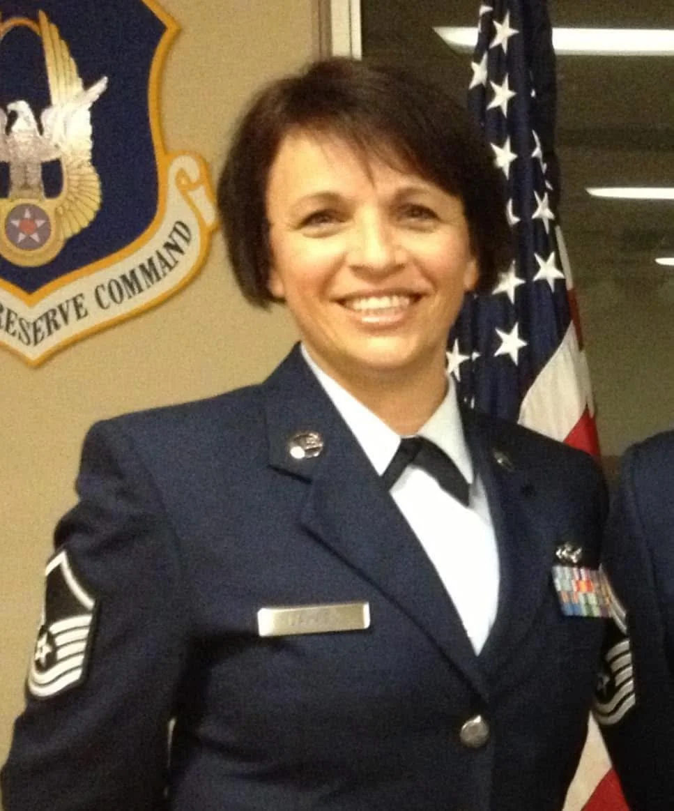 A female United States Air Force officer in dress uniform standing in front of an American flag and a Department of the Air Force insignia.