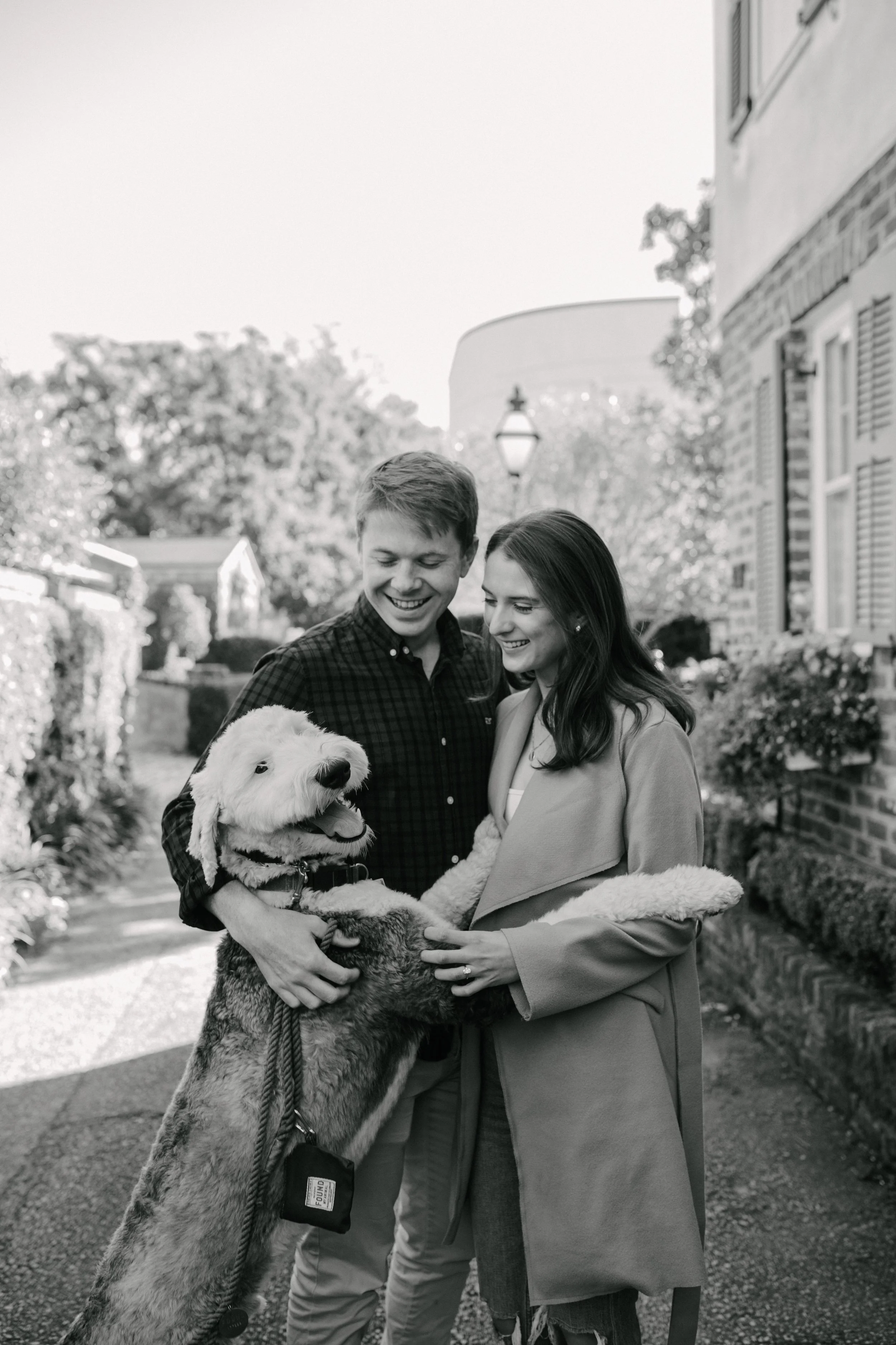 A smiling young man and woman are standing outside, holding a large, happy dog together. The man is wearing a checkered shirt and the woman is dressed in a coat. They appear to be enjoying a pleasant moment on a sunny day in a neighborhood with houses, trees, and a streetlamp in the background.