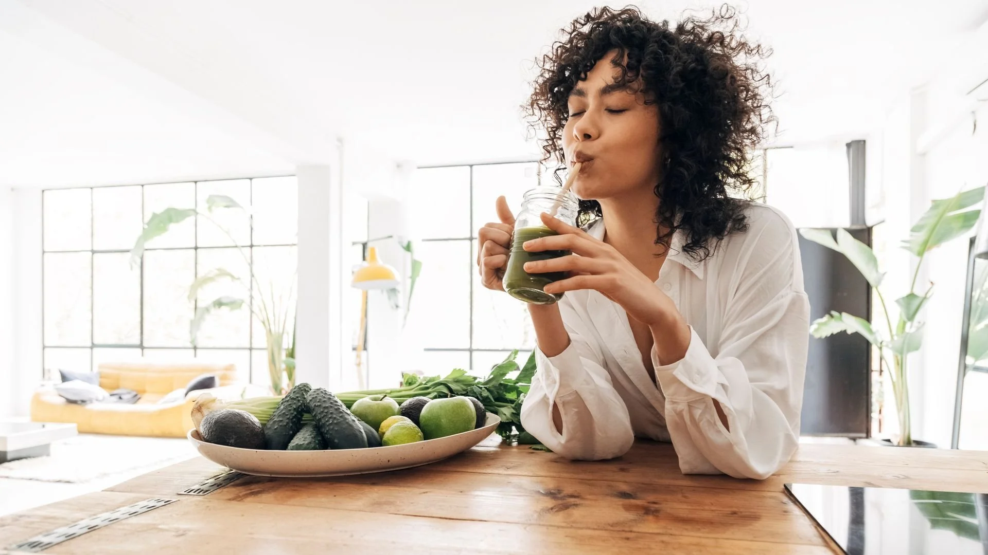 Woman drinking green smoothie with fruit and vegetables on table in bright room.