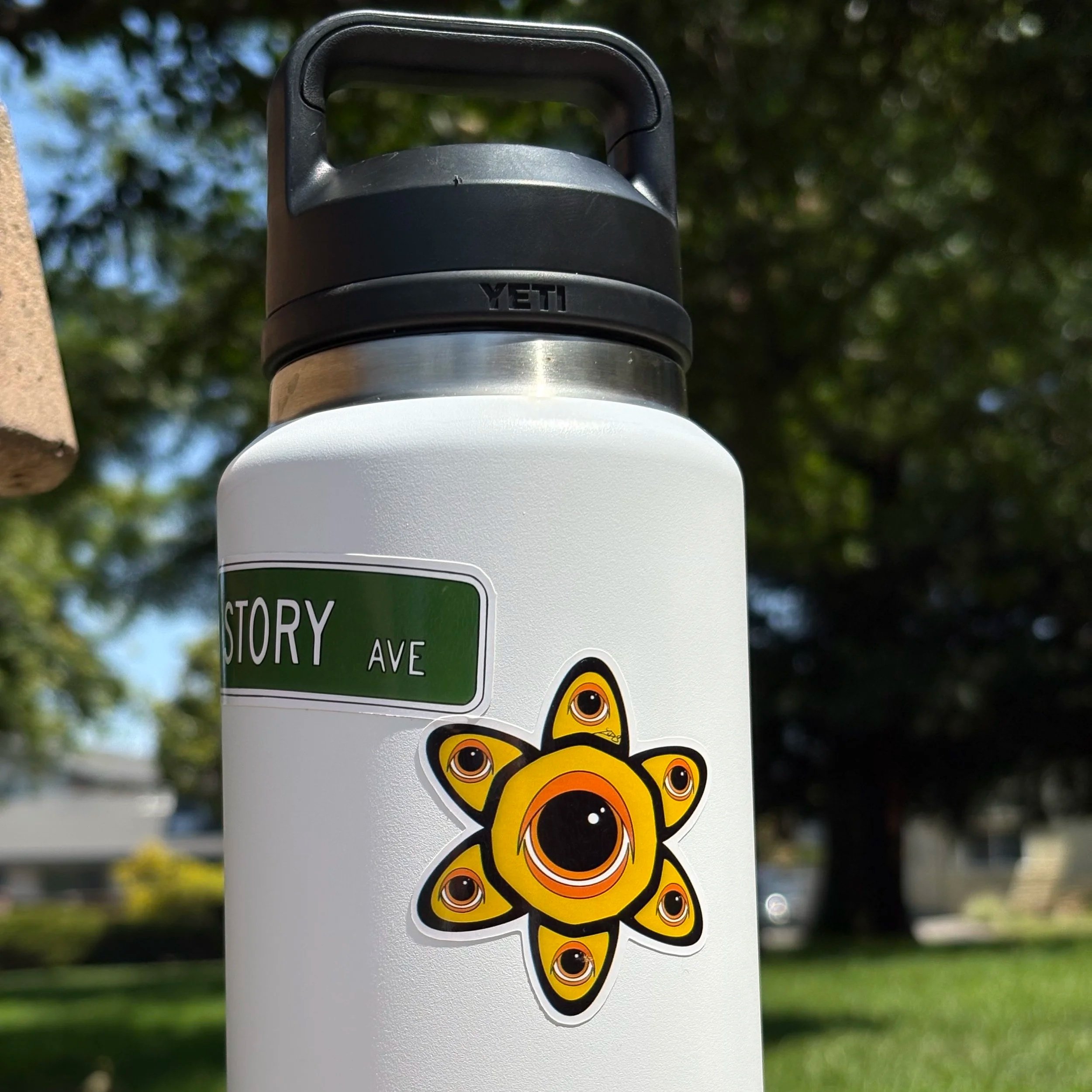 White water bottle with the green "STORY AVE" street sign sticker and a yellow eye/flower sticker applied, shot outdoors with trees blurred in the background. Paired on the page with a hoodie photo showing the same flower design applied to a black ho