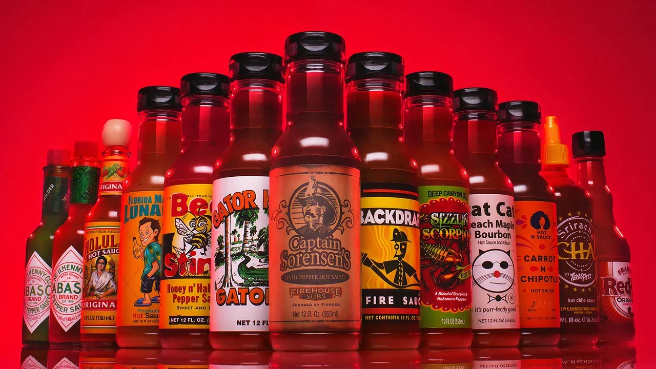 A collection of various hot sauce bottles with colorful labels, arranged on a red background.