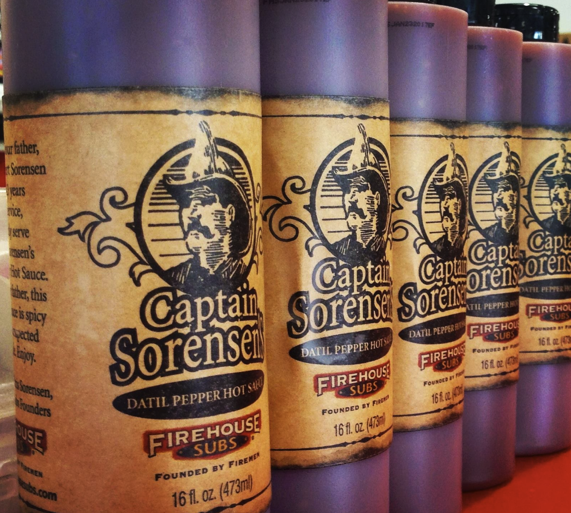 Cans of Captain Sorensen Datil Pepper Hot Sauce from Firehouse Subs, lined up on a red surface. The cans are purple with a paper label featuring a pirate's face and branding for Firehouse Subs.