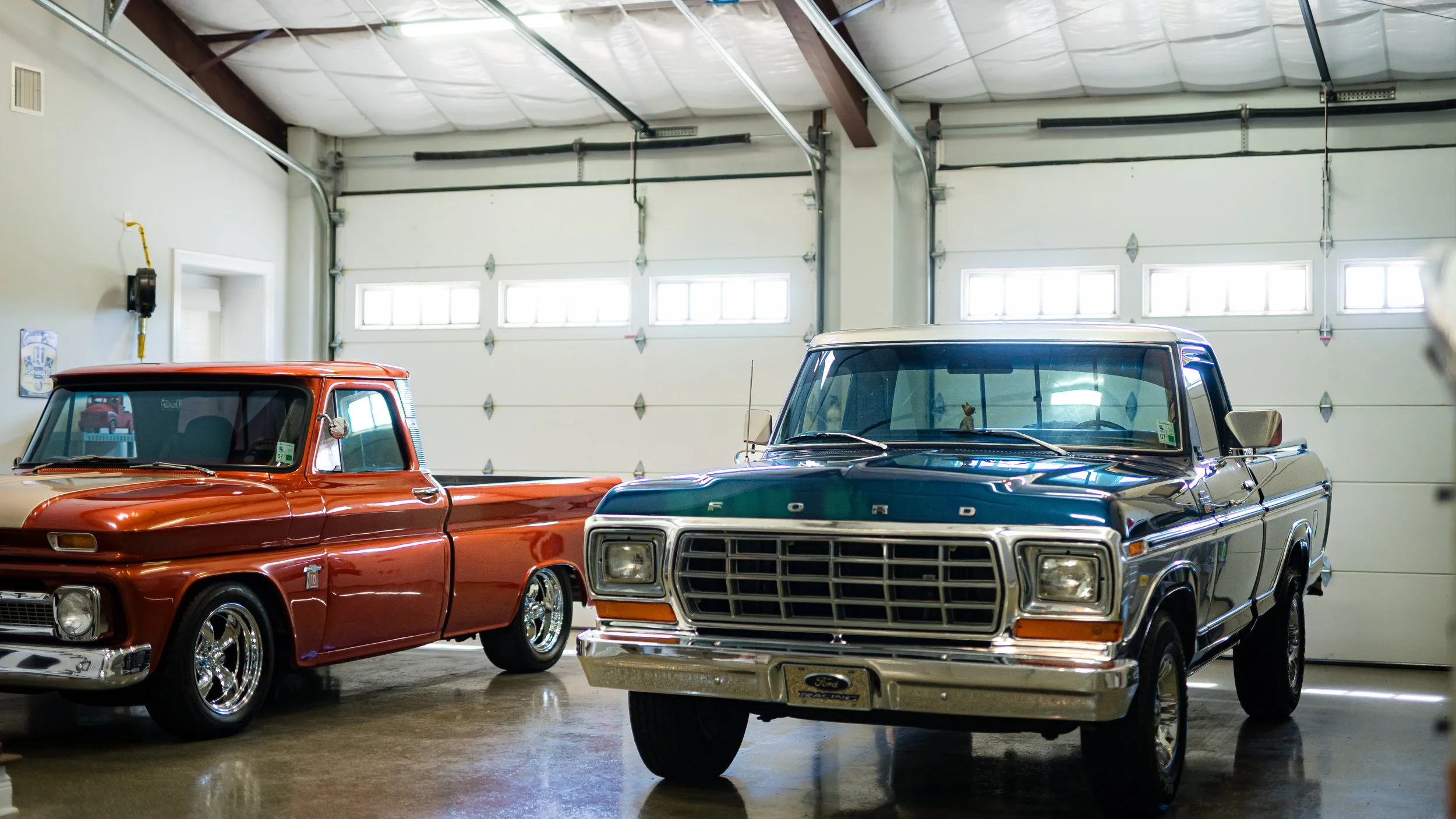 Two vintage pickup trucks, one red and one black, parked inside a garage with white walls and a high ceiling.