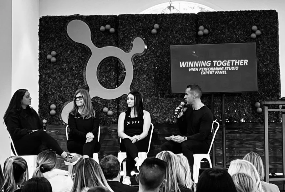 JJ Creegan leading a discussion on "Winning Together High Performing Studio Expert Panel." The backdrop features a large geometric design with a textured wall and decorative elements.