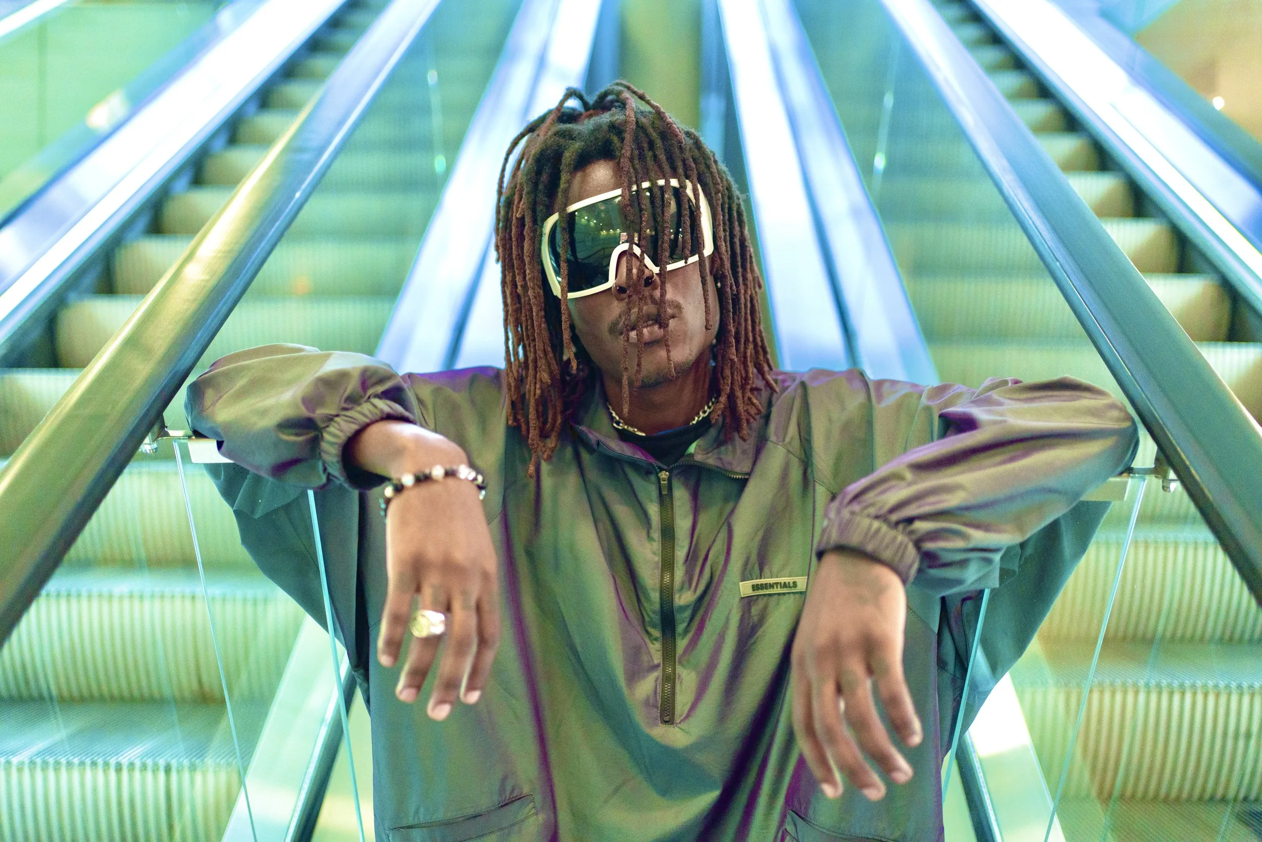 Young Gstar with dreadlocks wearing goggles/sunglasses and a Fear of God Essentials jacket, leaning on escalator handrails in a modern, brightly lit environment.