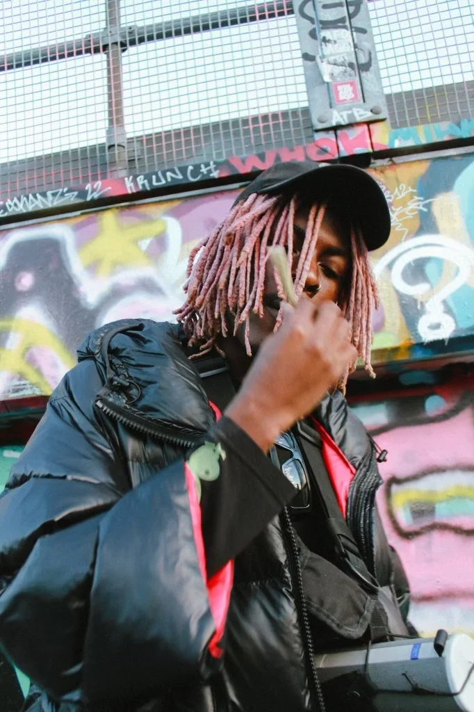 Young Gstar in a black cap and puffer jacket holding an object near their face, standing in front of a colorful graffiti wall.