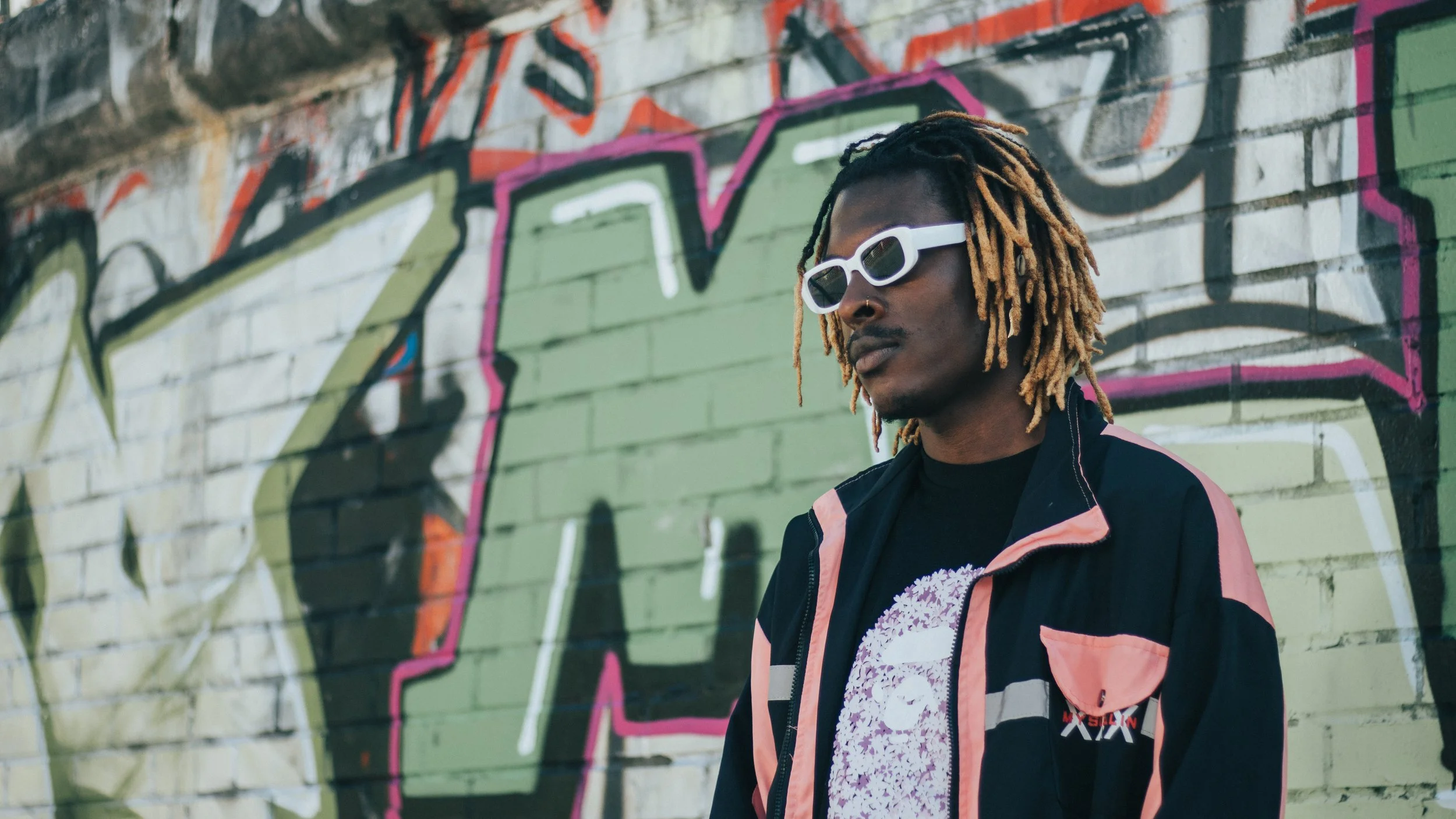 Young Gstar with blonde dreadlocks and white sunglasses standing in front of a graffiti-covered wall.