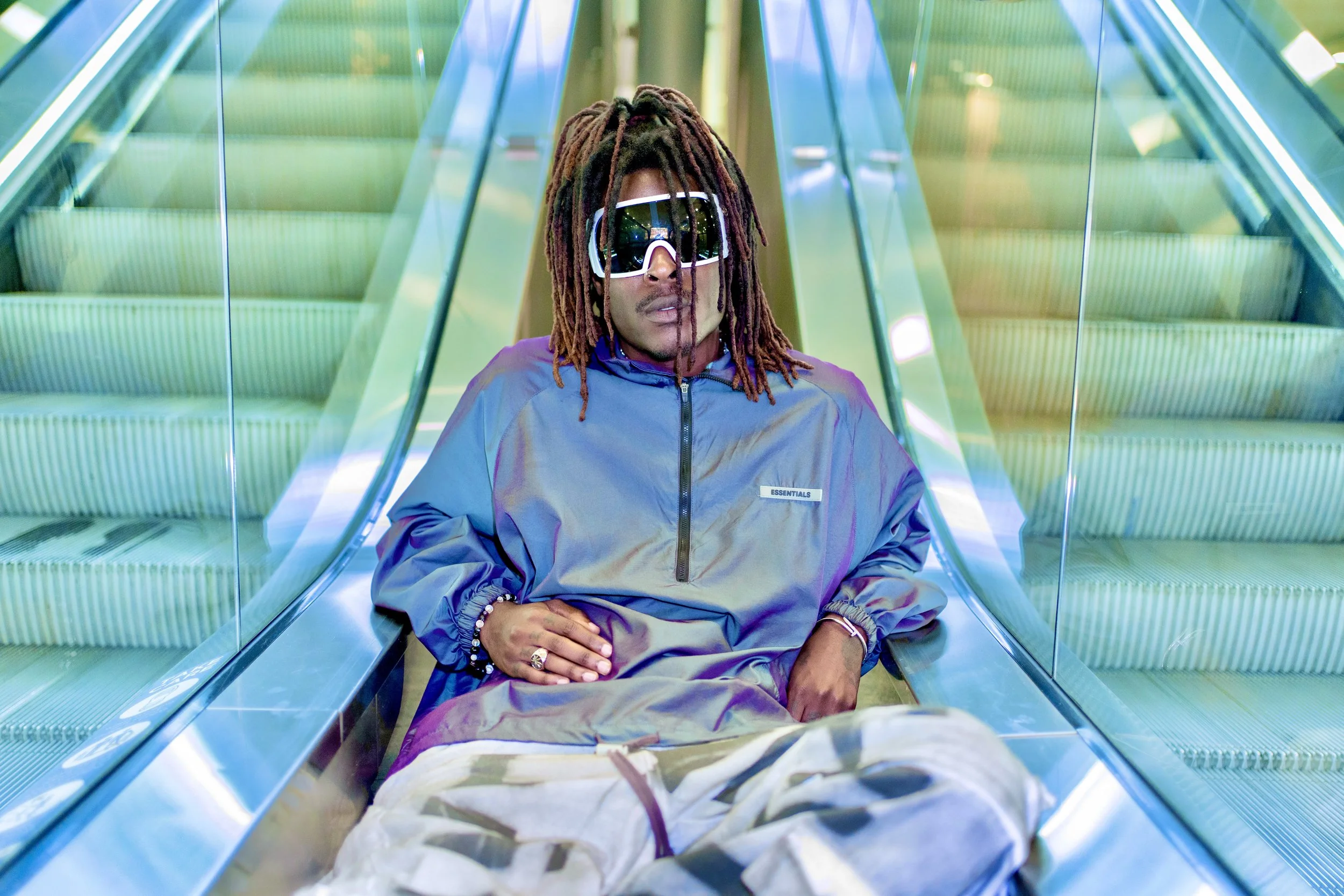 Young Gstar with dreadlocks wearing sunglasses and a metallic a Fear of God Essentials jacket sitting on the escalator in a modern setting.