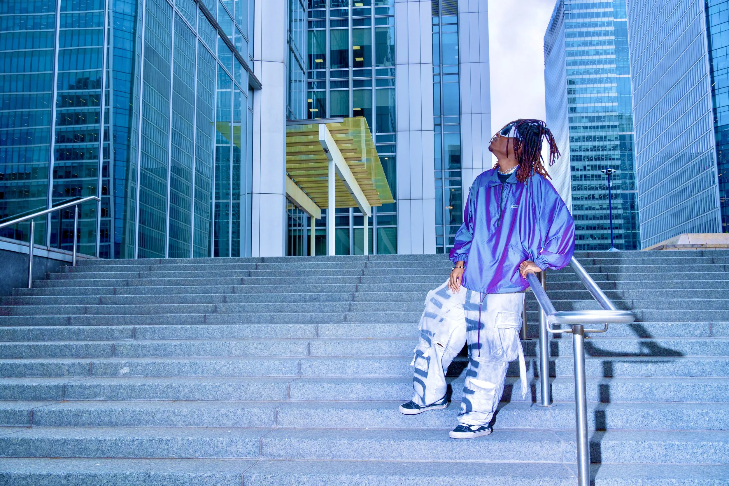Young Gstar with long dreadlocks wearing a shiny purple jacket and baggy cargo pants standing on city steps near modern glass skyscrapers.
