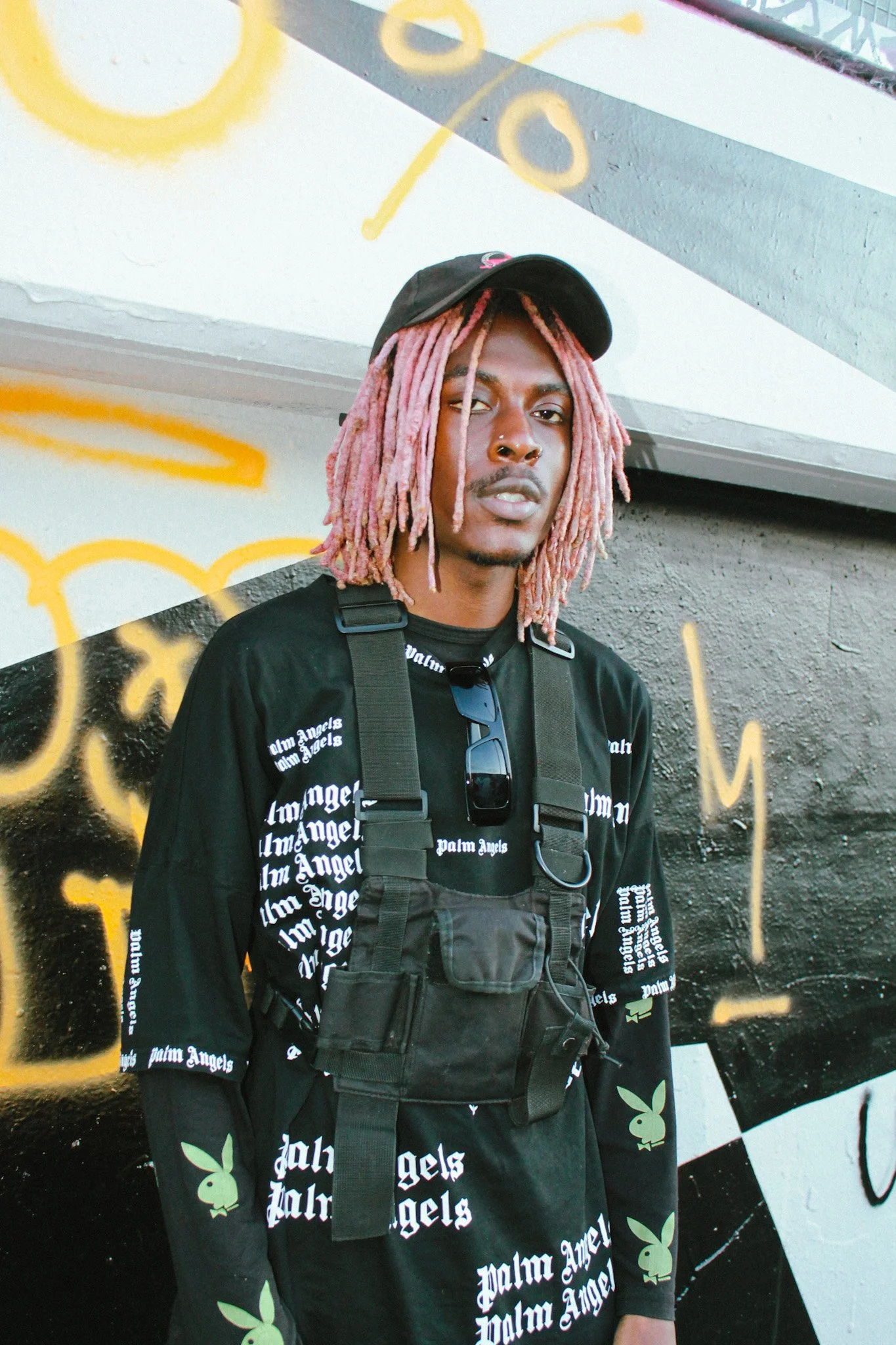 Young Gstar with pink dreadlocks wearing a black cap, sunglasses, and a black shirt with white text and graphics, standing against a graffiti-covered wall.