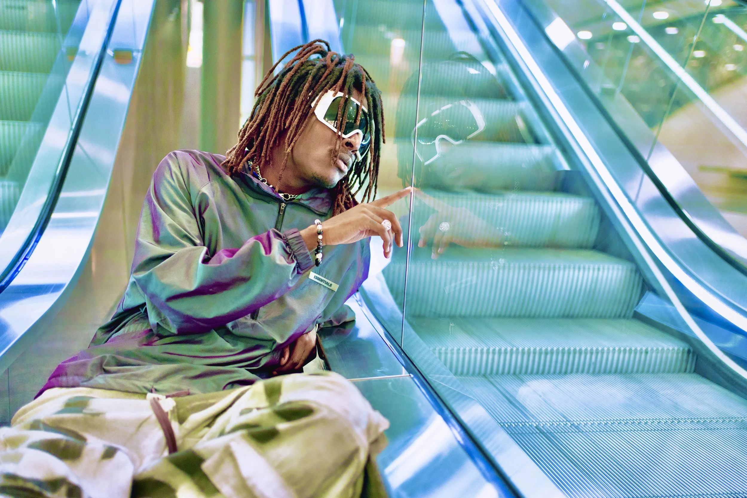 Young Gstar with dreadlocks wearing sunglasses, a jacket, and white baggy pants, sitting on the floor beside an escalator, touching the glass railing.