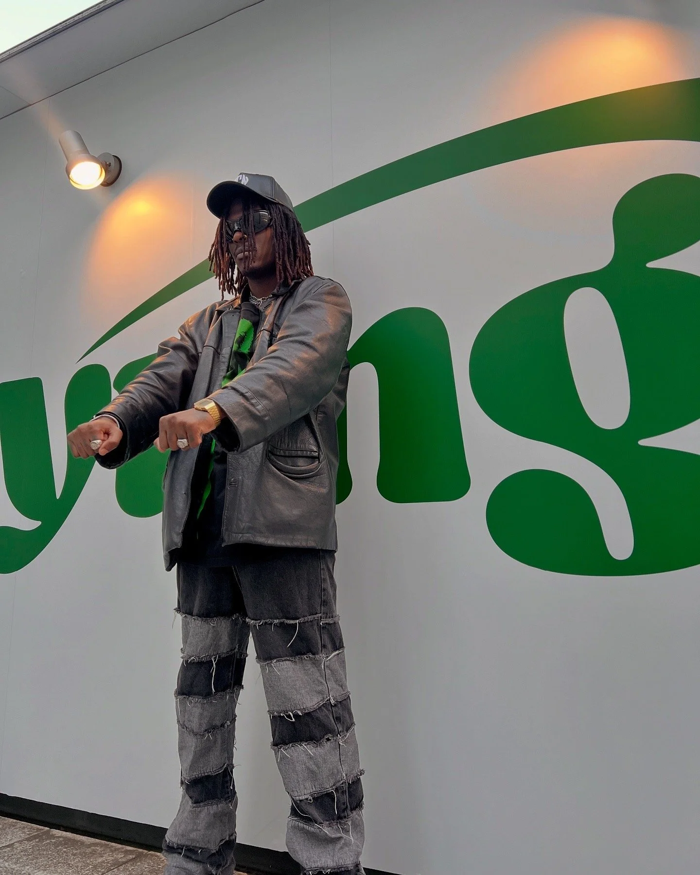 Young Gstar wearing a leather jacket, black cap, and patchwork jeans stands in front of a wall with green lettering.