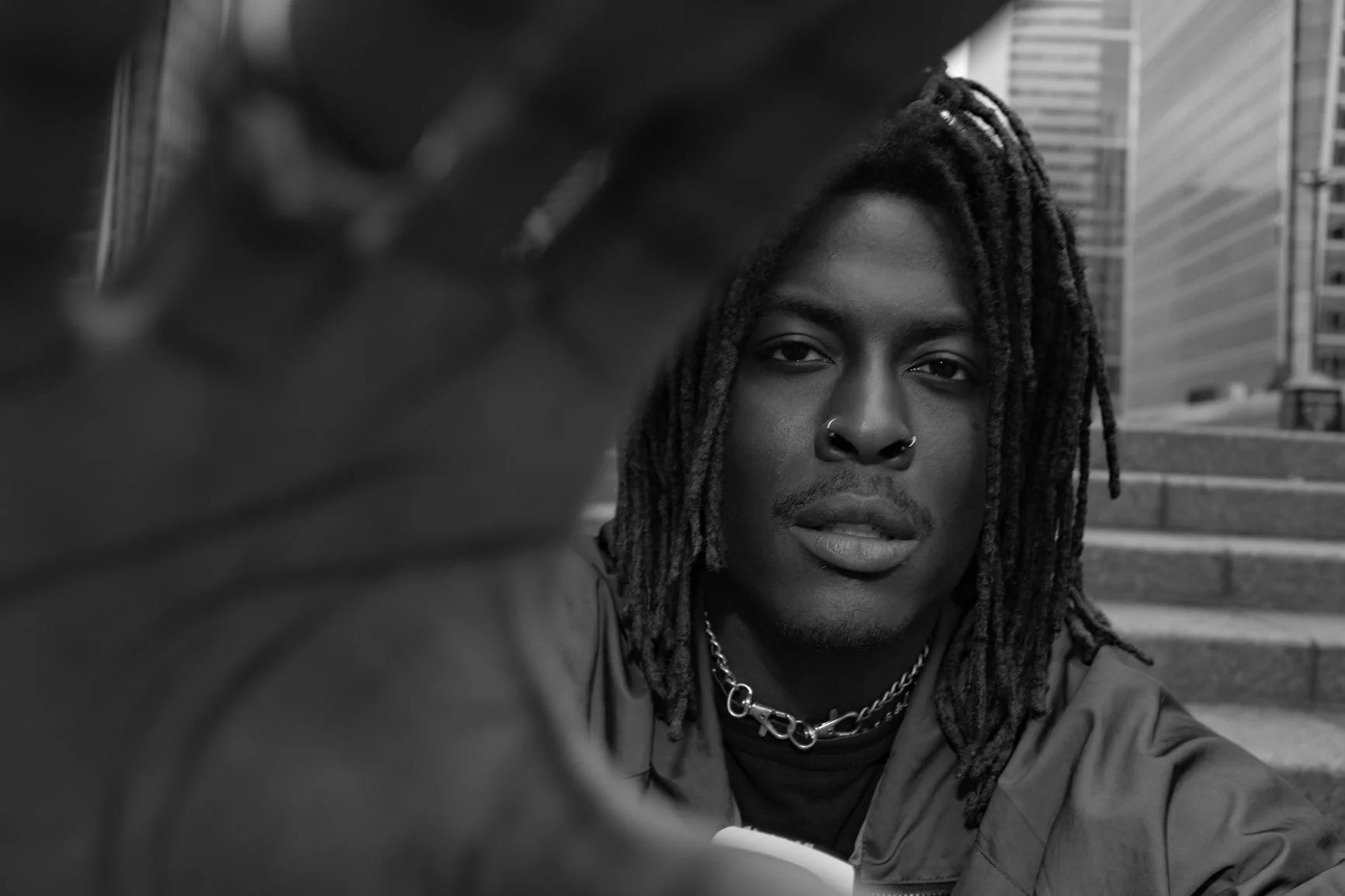 Black and white photo of Young Gstar with dreadlocks outdoors in an urban setting, with city buildings and stairs in the background.