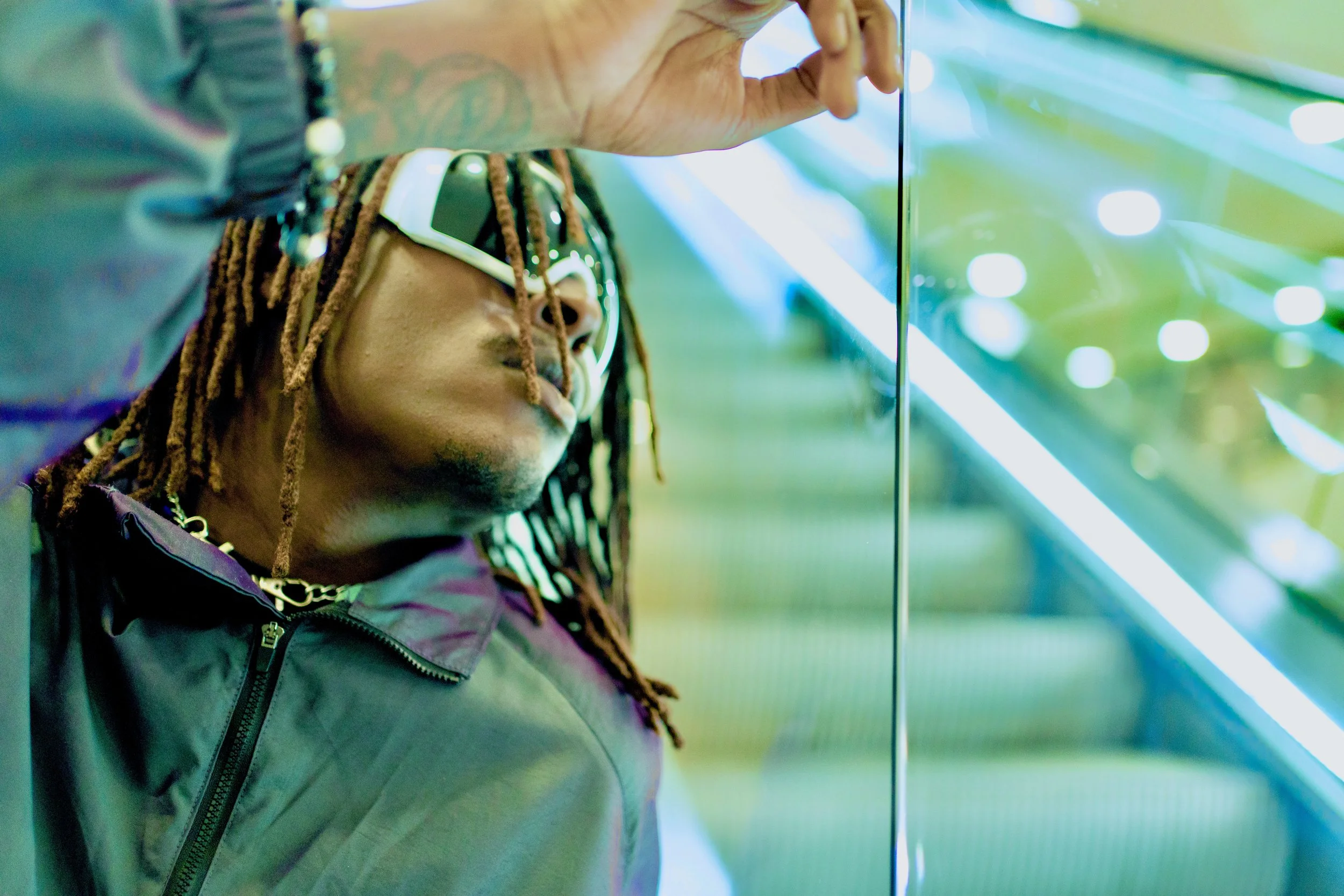 Young Gstar with dreadlocks, sunglasses, and a jacket, reflected in a glass surface, with neon lights in the background.