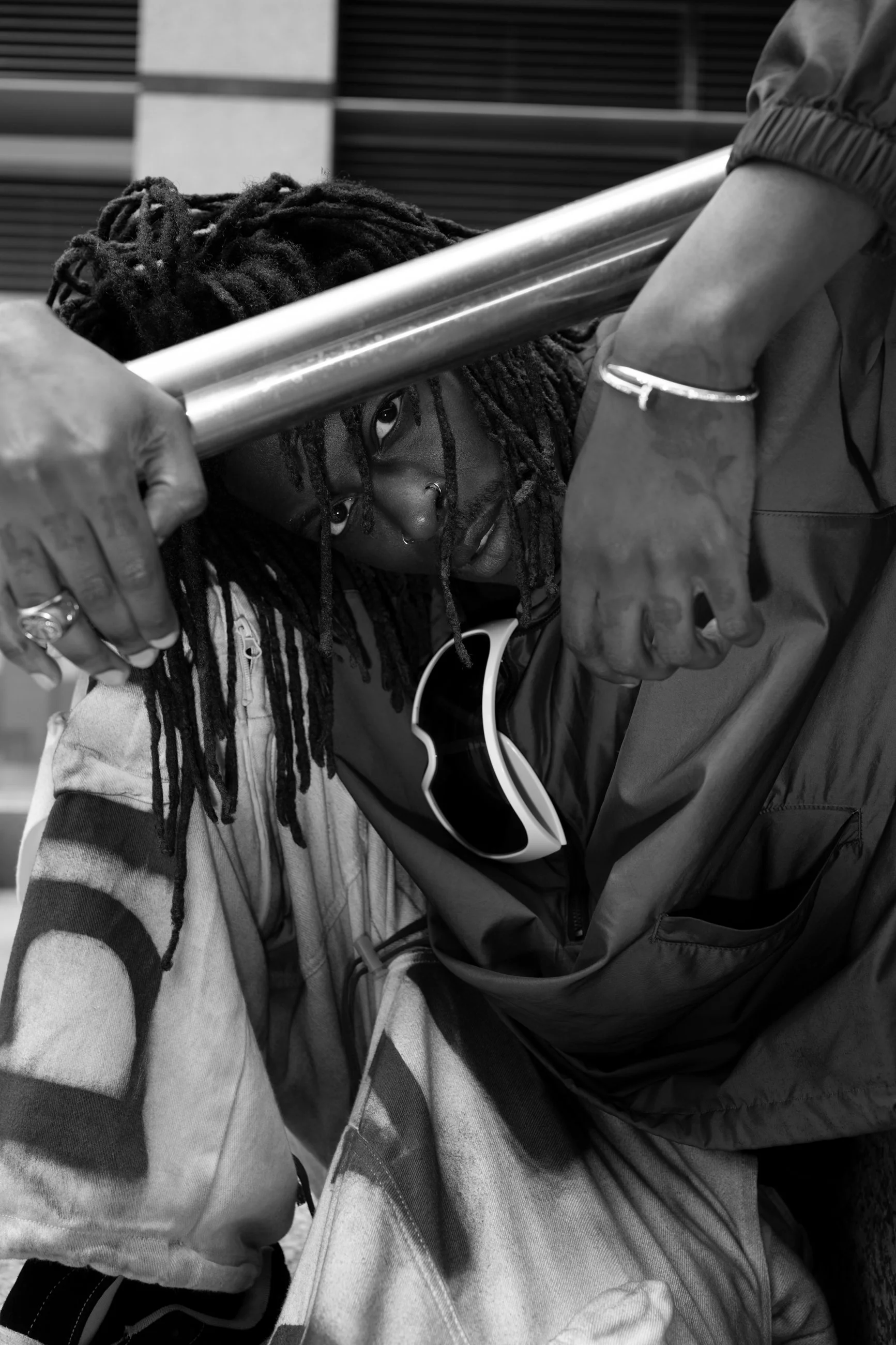 Young Gstar with dreadlocks leaning their head through the gap between two metal bars, looking directly at the camera, with sunglasses hanging from their shirt. Part of an additional person wearing a jacket and rings is visible, holding the bars.