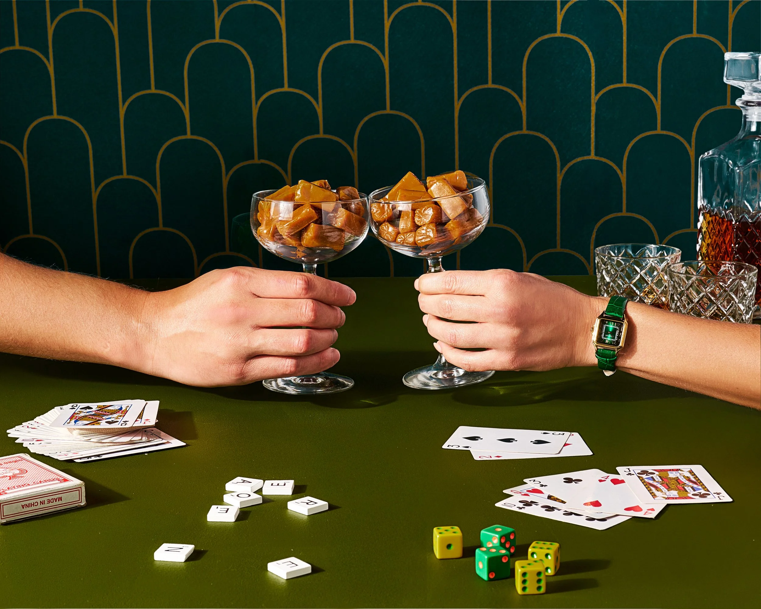 An elegant photo of two hands holding goblets with caramels in them doing a cheers, surrounded by playing cards and dice on a art deco inspired green background.