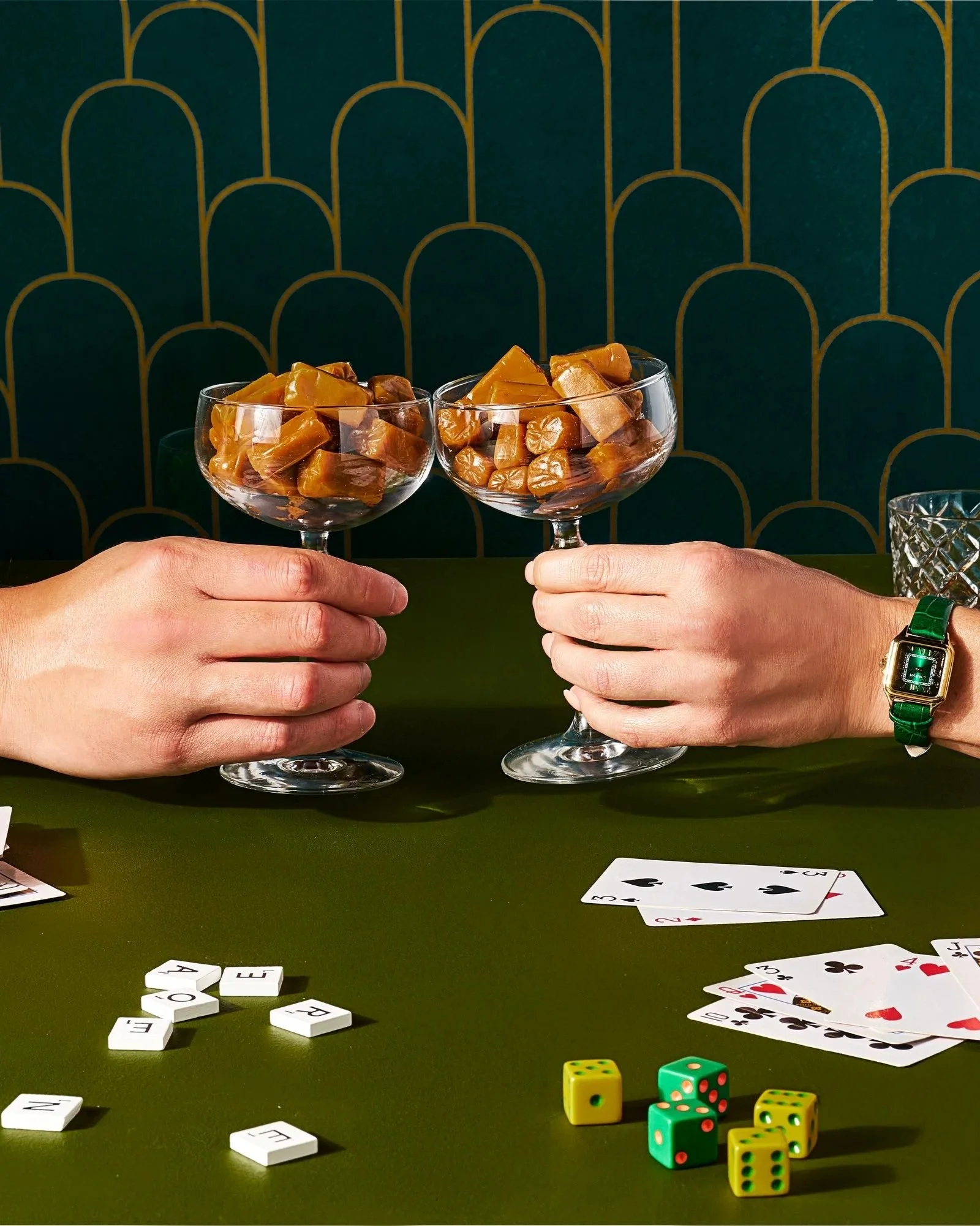 An elegant photo of two hands holding goblets with caramels in them doing a cheers, surrounded by playing cards and dice on a art deco inspired green background. 