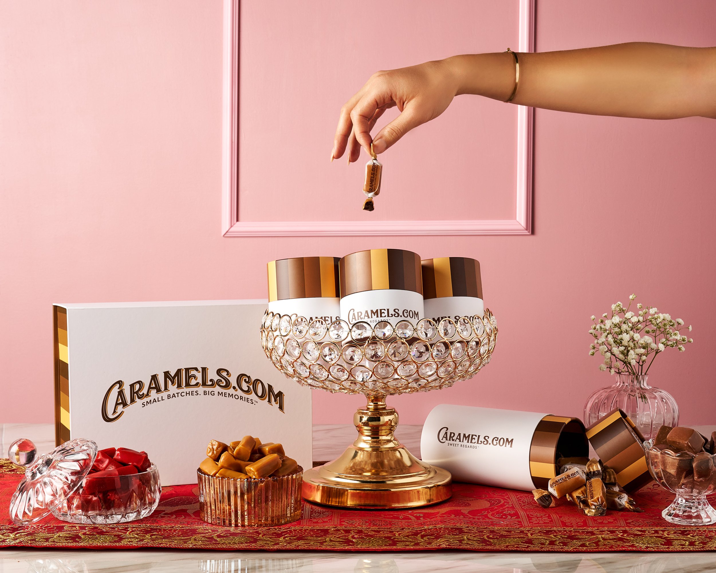 Elegant product photo of Caramels.com gourmet treats styled with luxury gold and crystal accents on a bright pink background surrounded by different candy bowls and box sets.