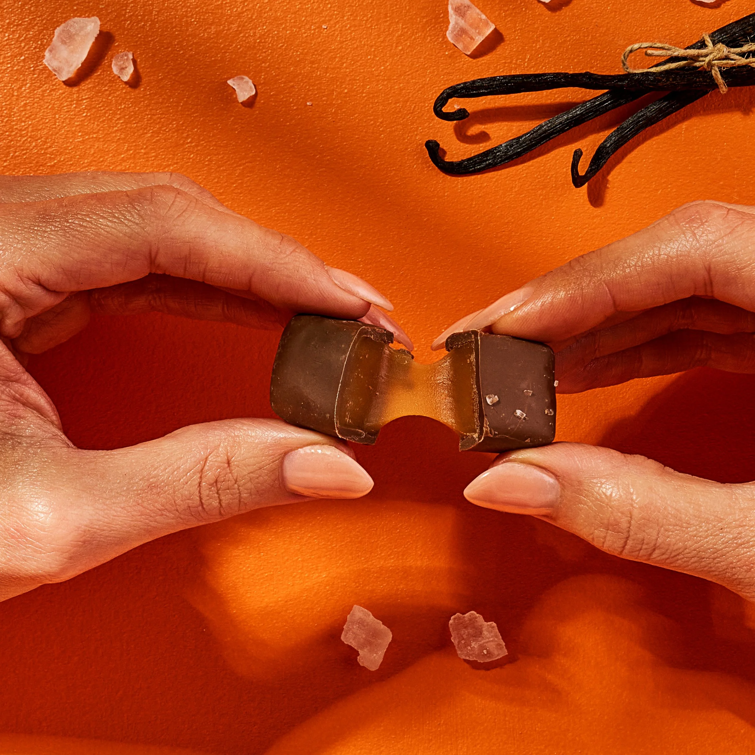 A womans hands gently pull apart a chocolate square with caramel filling as it oozes out on a bright orange backdrop. 