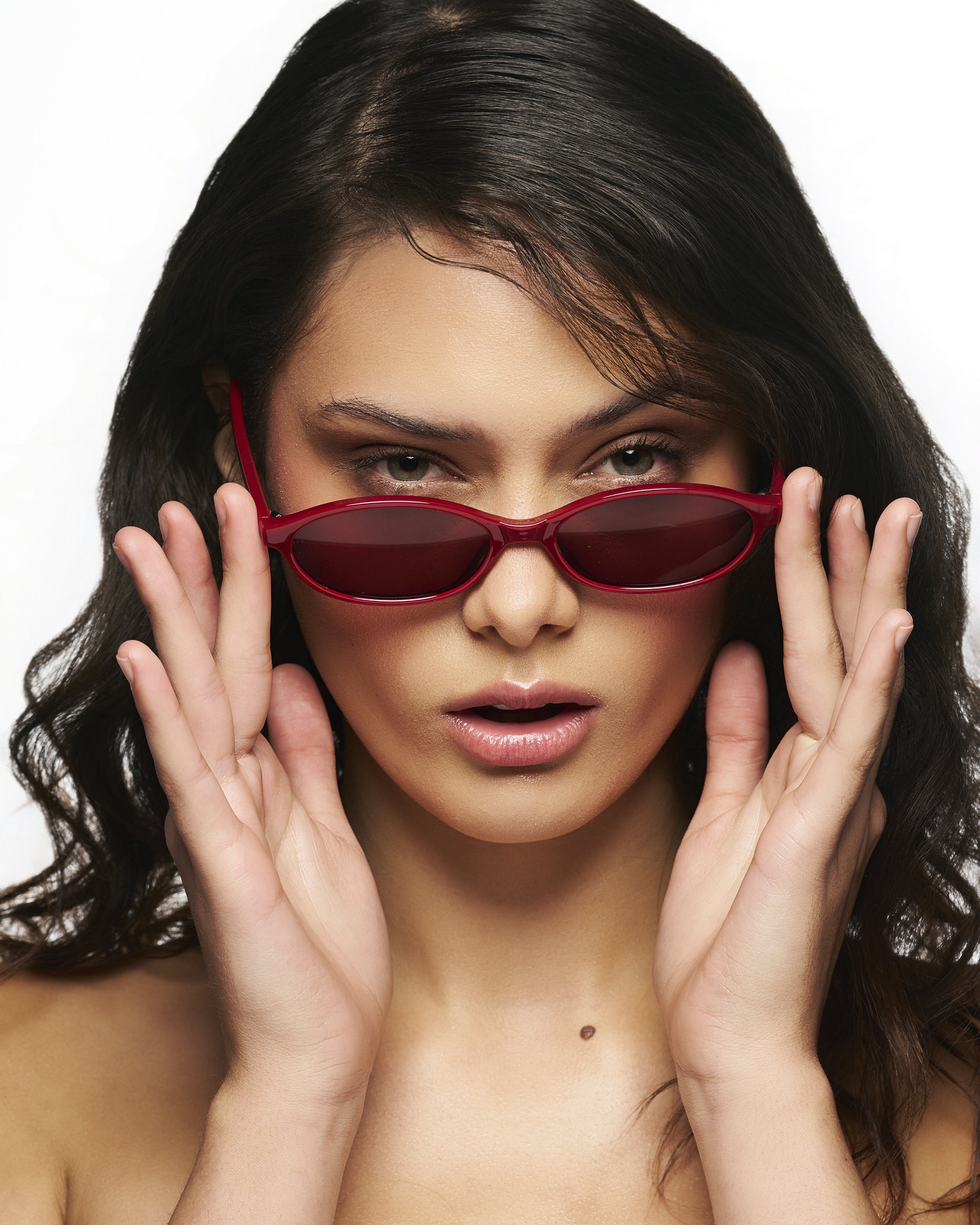 A stylish portrait of a woman showcasing shades with a red tint. She holds each side while sliding it down her nose. Perfect for editorial campaigns. 