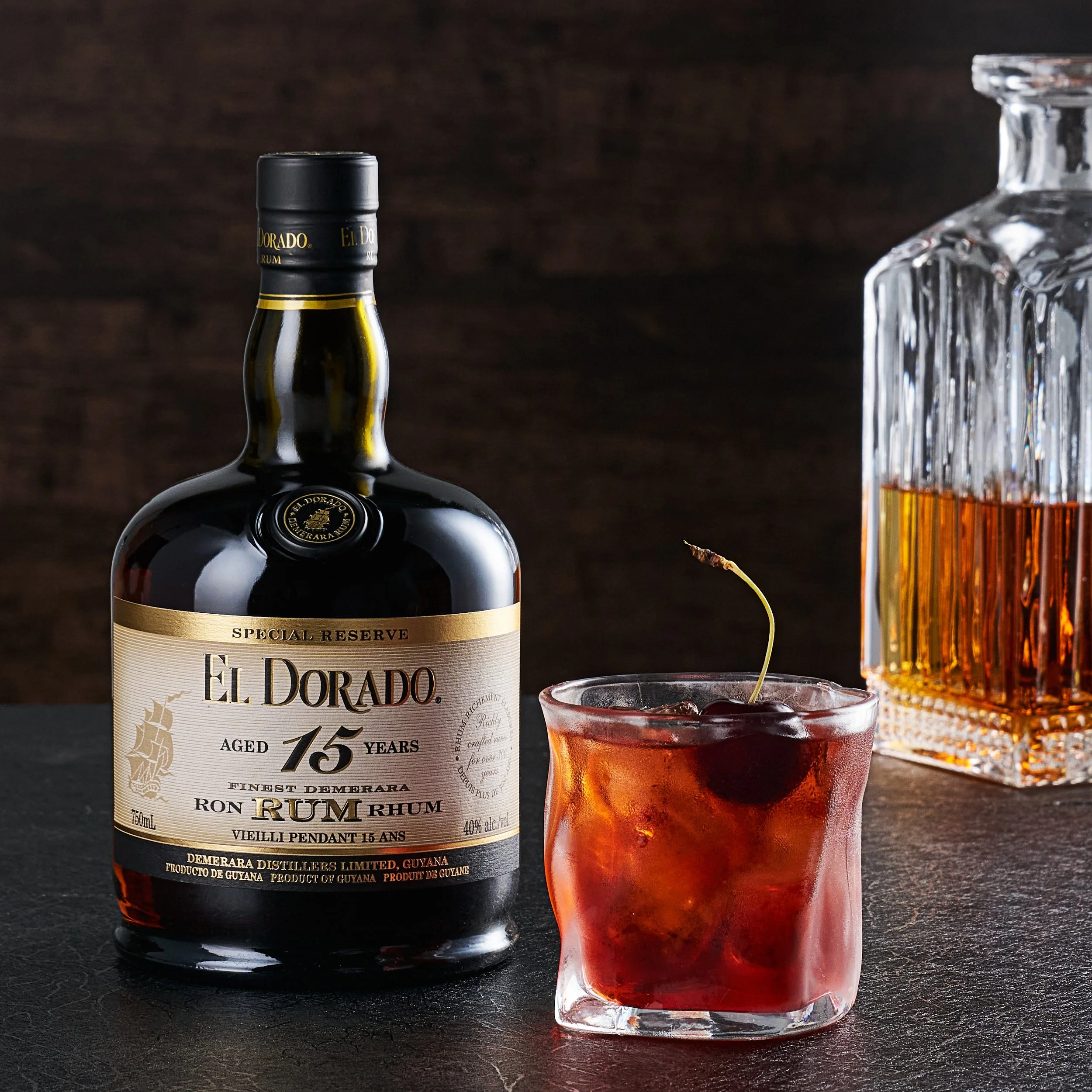 Photograph of a dark wooden table with a bottle of El Dorado Special Reserve Rum, aged 15 years, and a glass of dark rum with a cherry garnish. Beside the bottle is a glass decanter of amber liquid. The background is a textured dark brown.