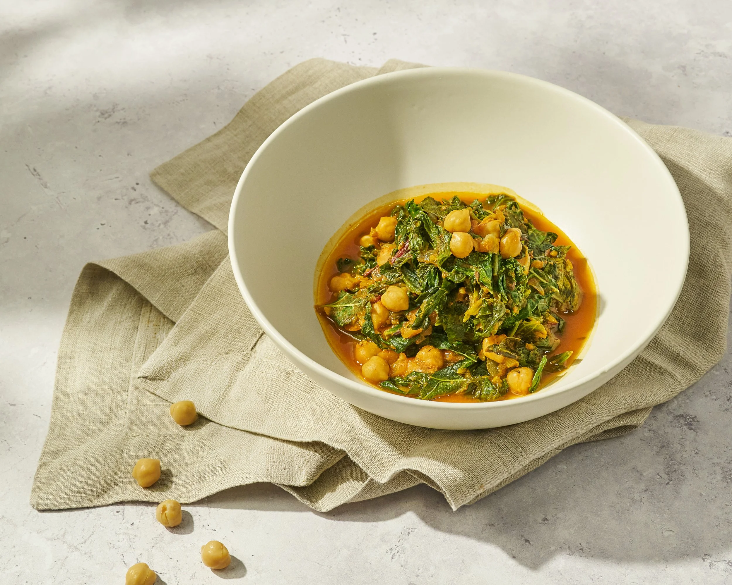 A white bowl filled with chickpea and spinach curry, placed on a beige napkin on a gray surface with a few chickpeas scattered around.