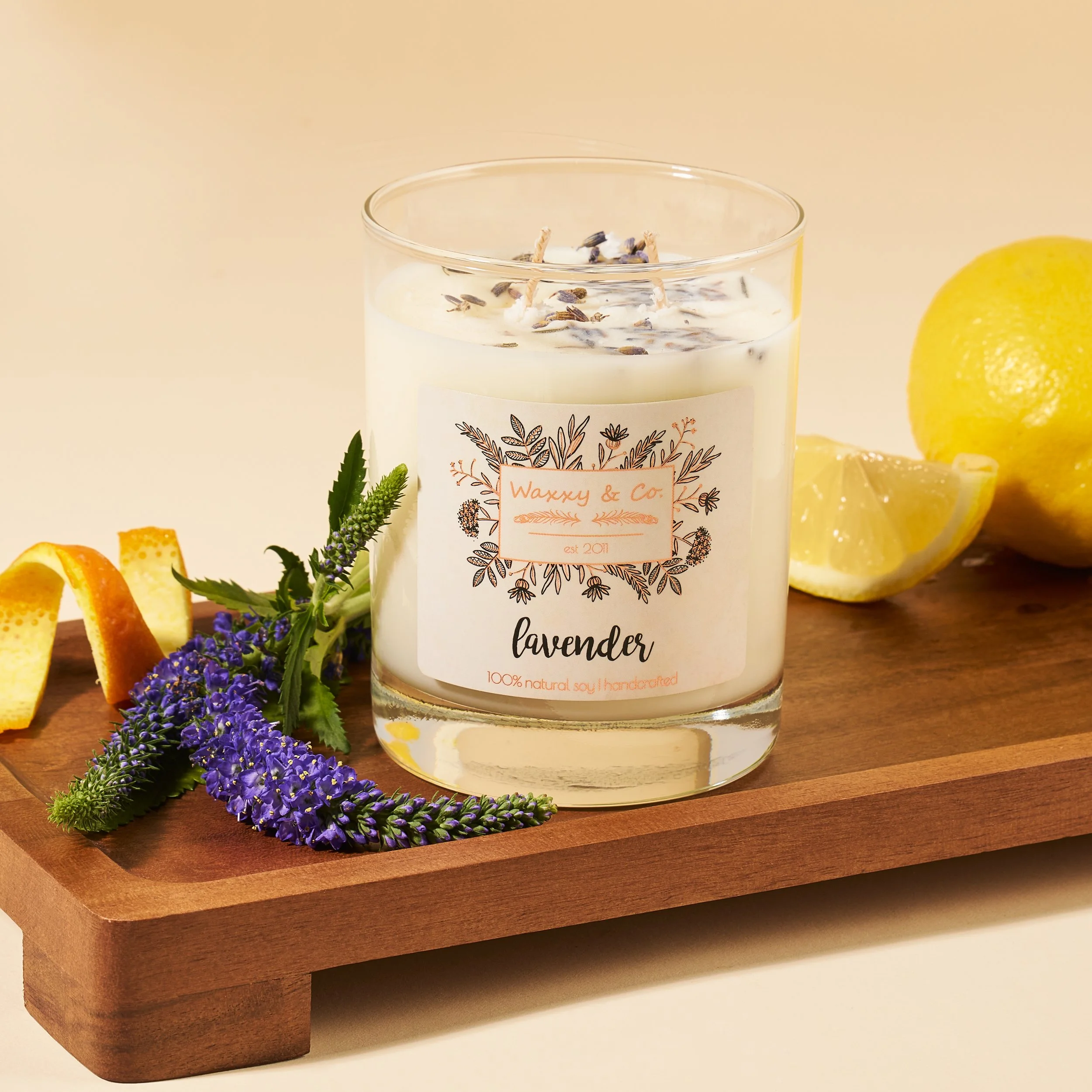 Lavender-scented candle on wooden board with purple flowers, lemon, and orange peel.